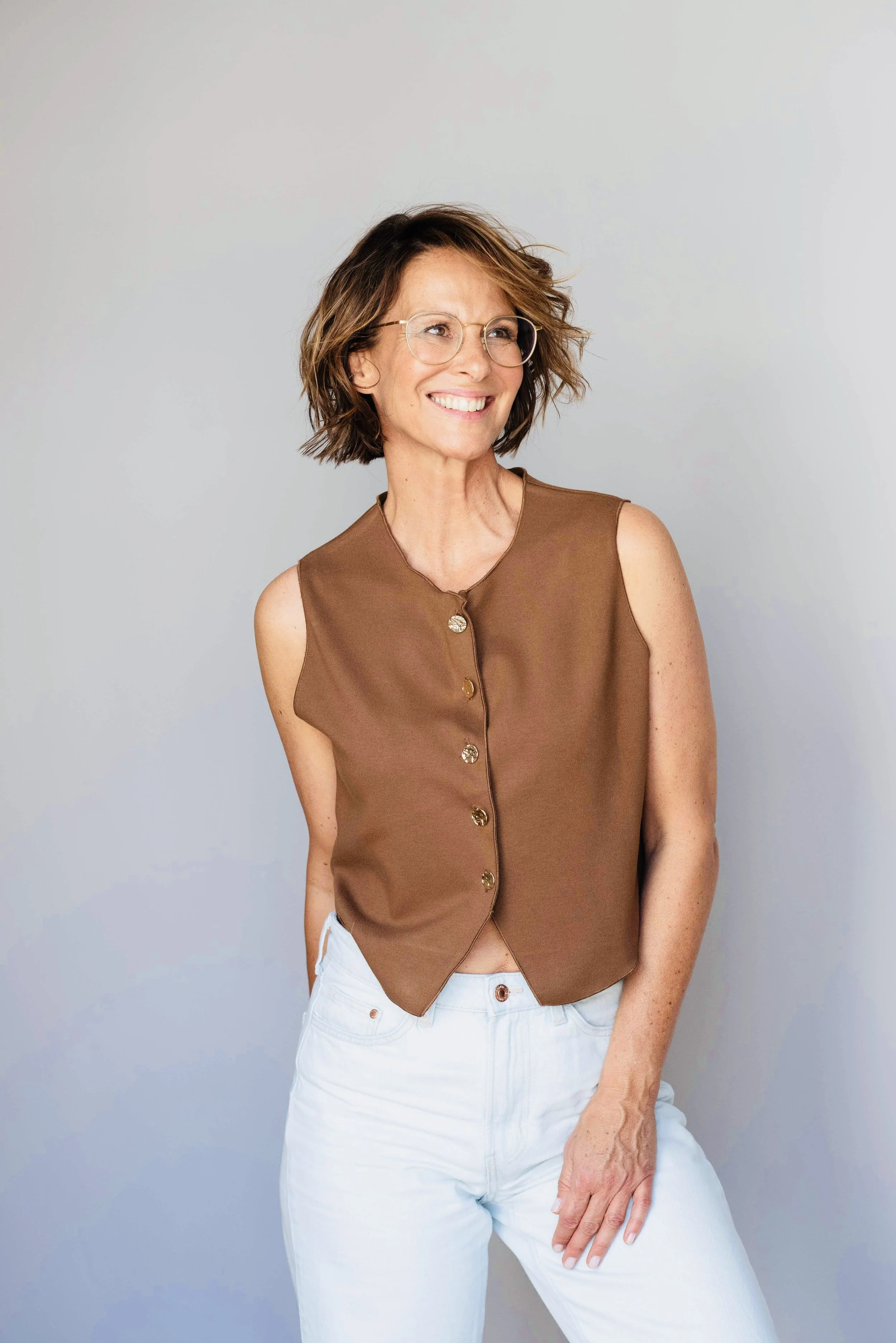 Lächelnde Frau mit Brille, kurze braune Locken, braunes ärmelloses Top, weiße Jeans vor neutralem Hintergrund.