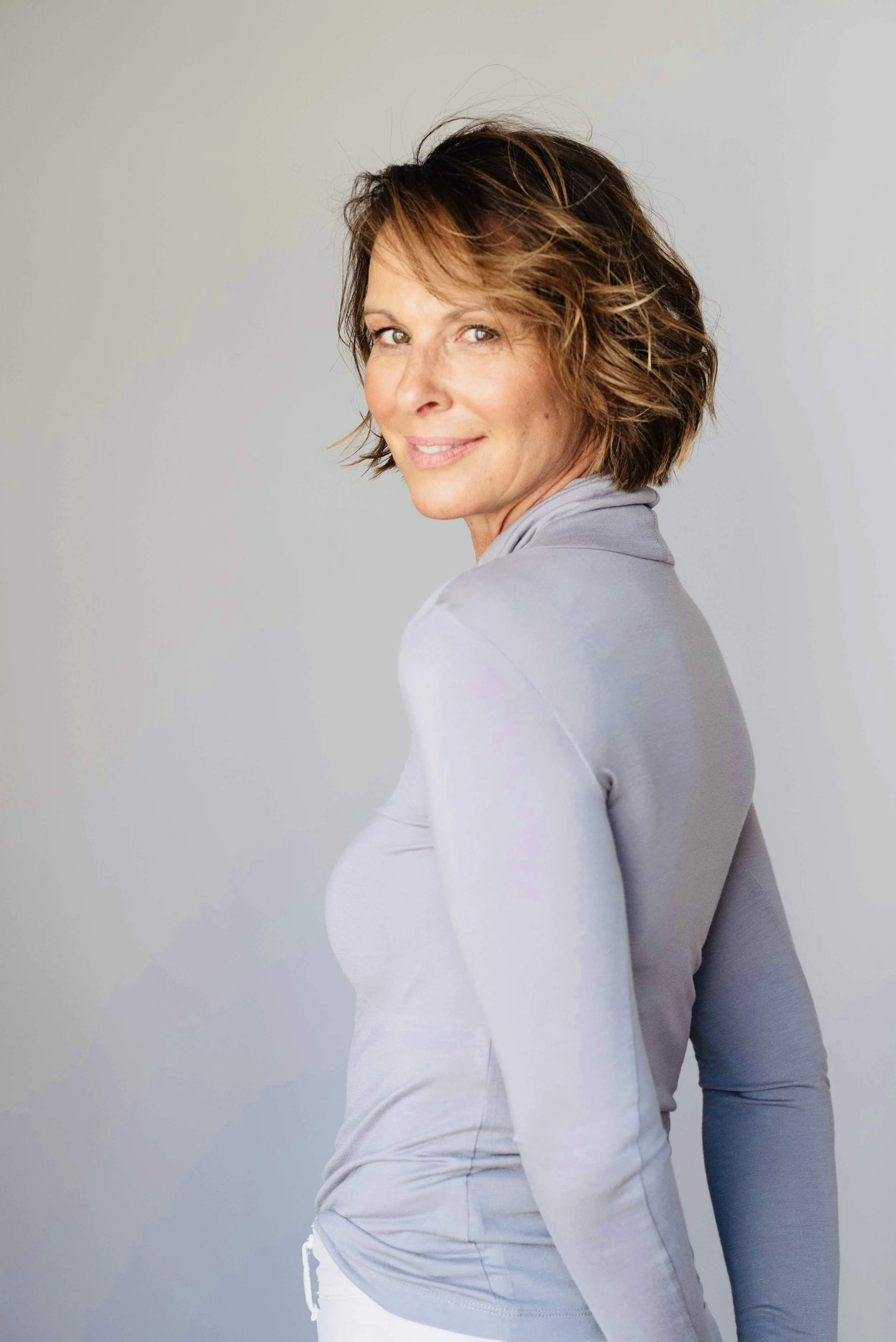 Eine lächelnde Frau mit kurzen braunen Haaren vor einem hellgrauen Hintergrund, trägt einen grauen langen Arm Pullover.