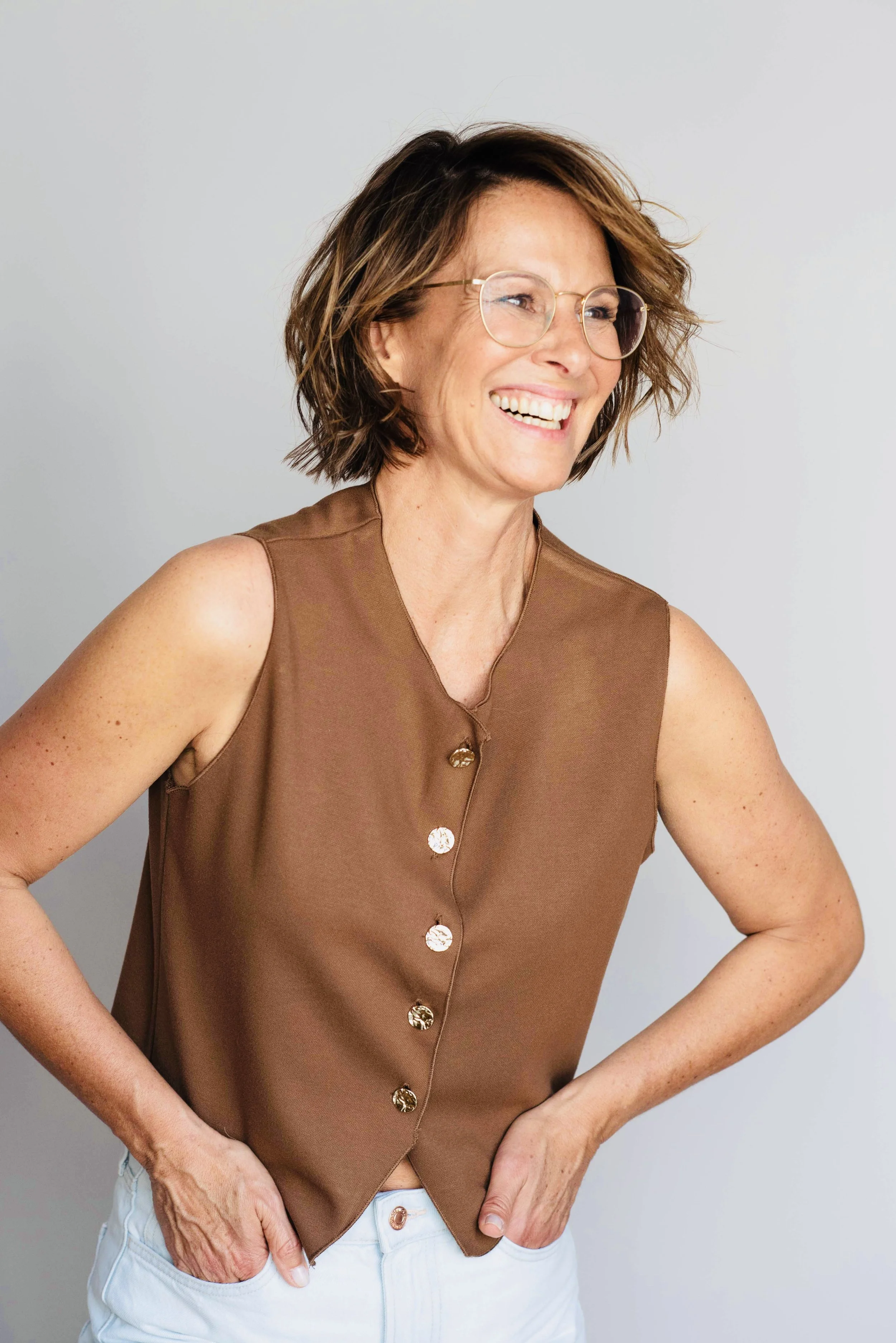 Eine lachende Frau mit kurz braunem Haar, Brille und braunem Top, die mit den Händen in den Taschen ihrer weißen Hose posiert.