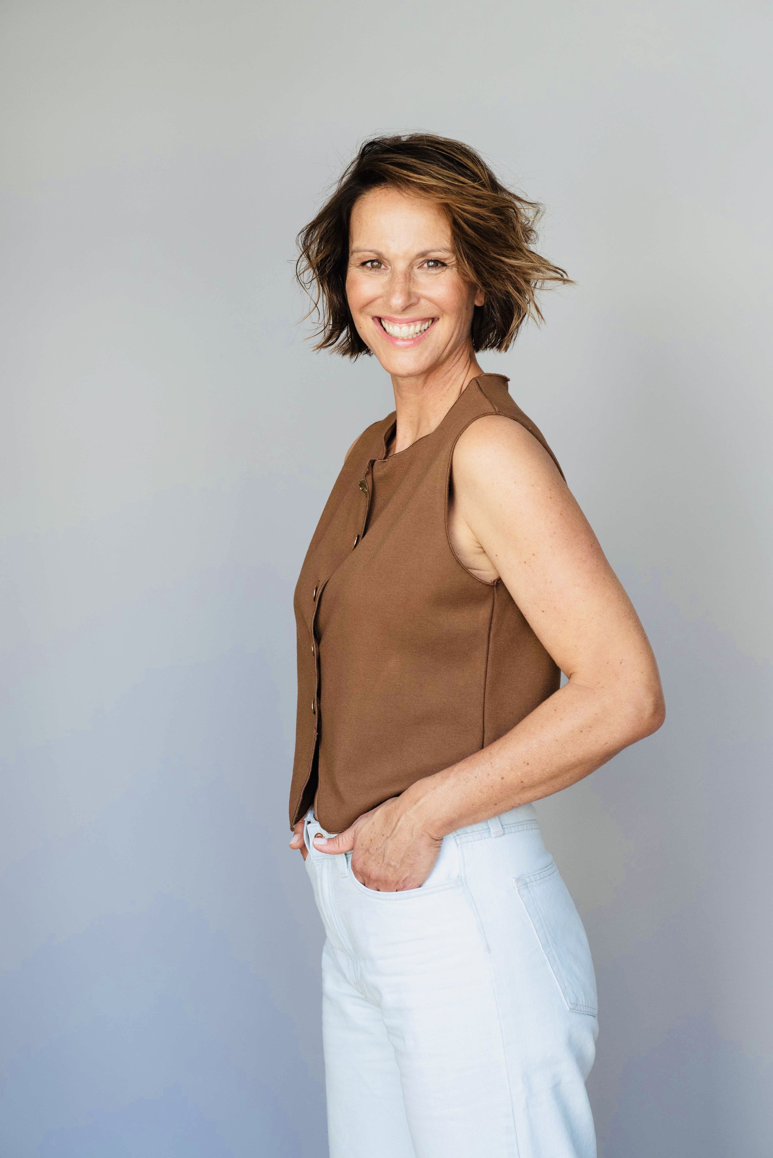 Eine lachende Frau mit kurzen braunen Haaren, trägt ein braunes Top und weiße Hosen, stehend vor neutralem Hintergrund.