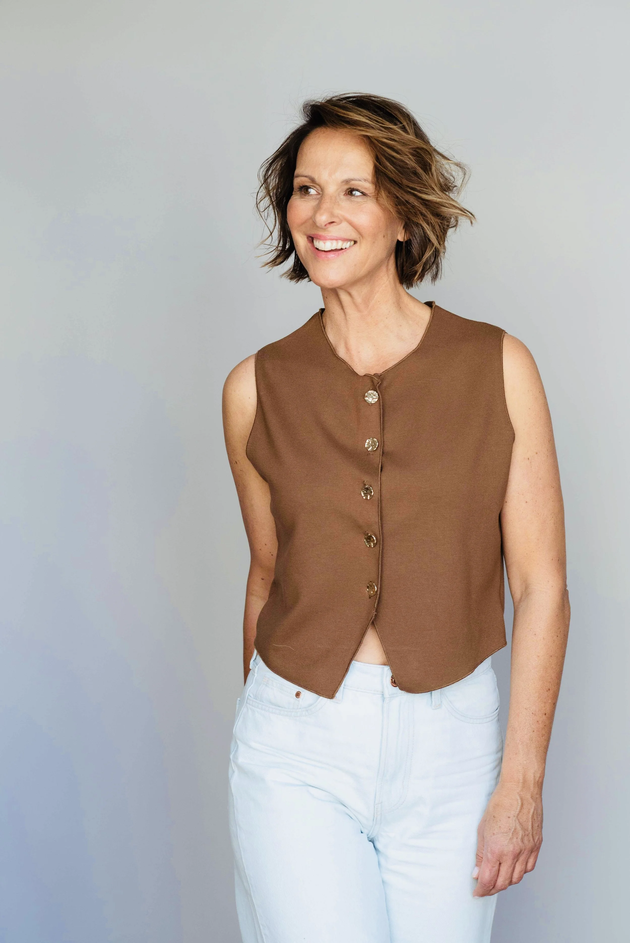 Eine lächelnde Frau mit kurzen, braunen, gewellten Haaren trägt eine braune ärmellose Jacke und weiße Jeans vor einem hellgrauen Hintergrund.