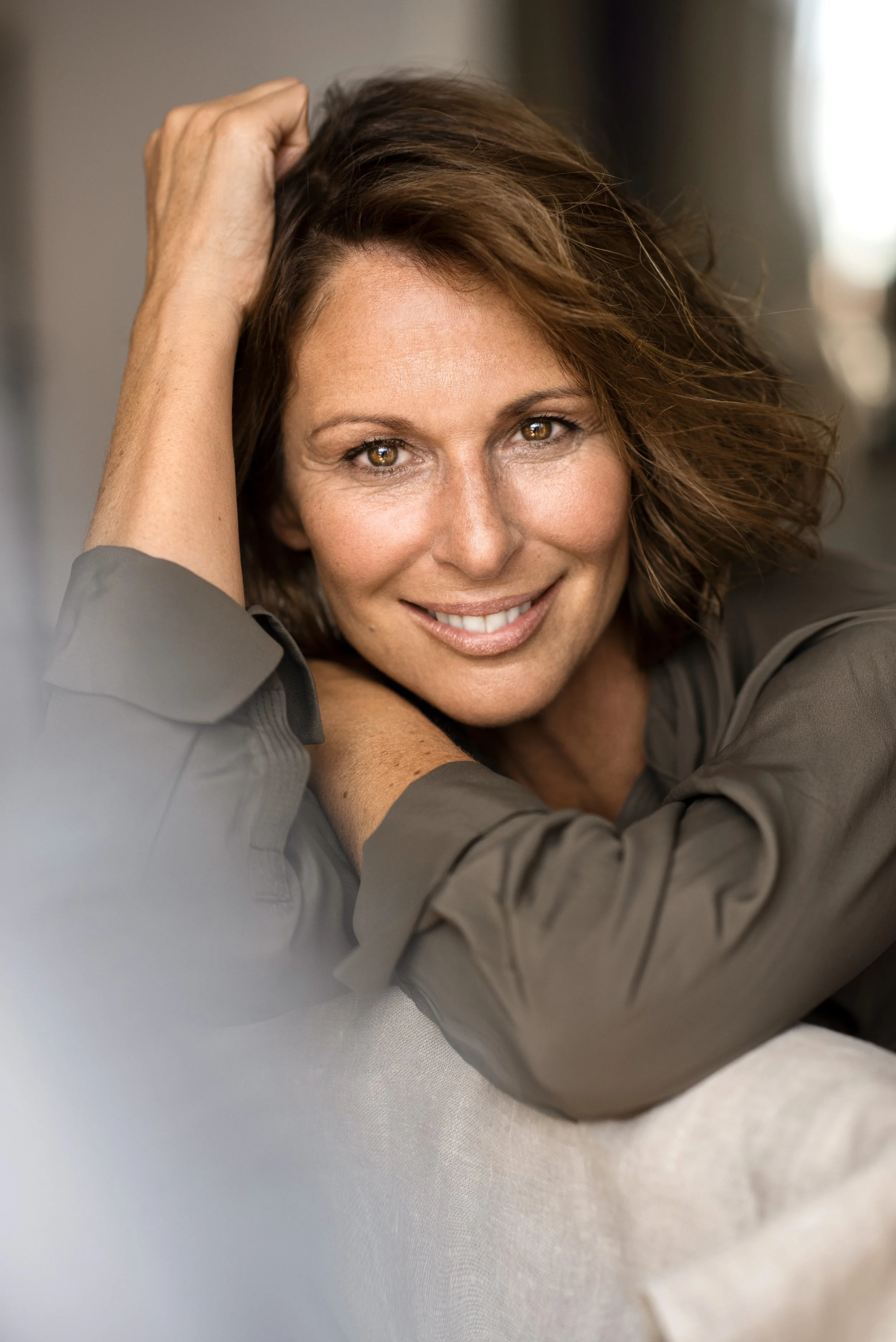 Nahaufnahme einer lachenden Frau mit braunen, lockeren Haaren, die auf einem Sofa liegt, mit einem freundlichen Gesichtsausdruck.