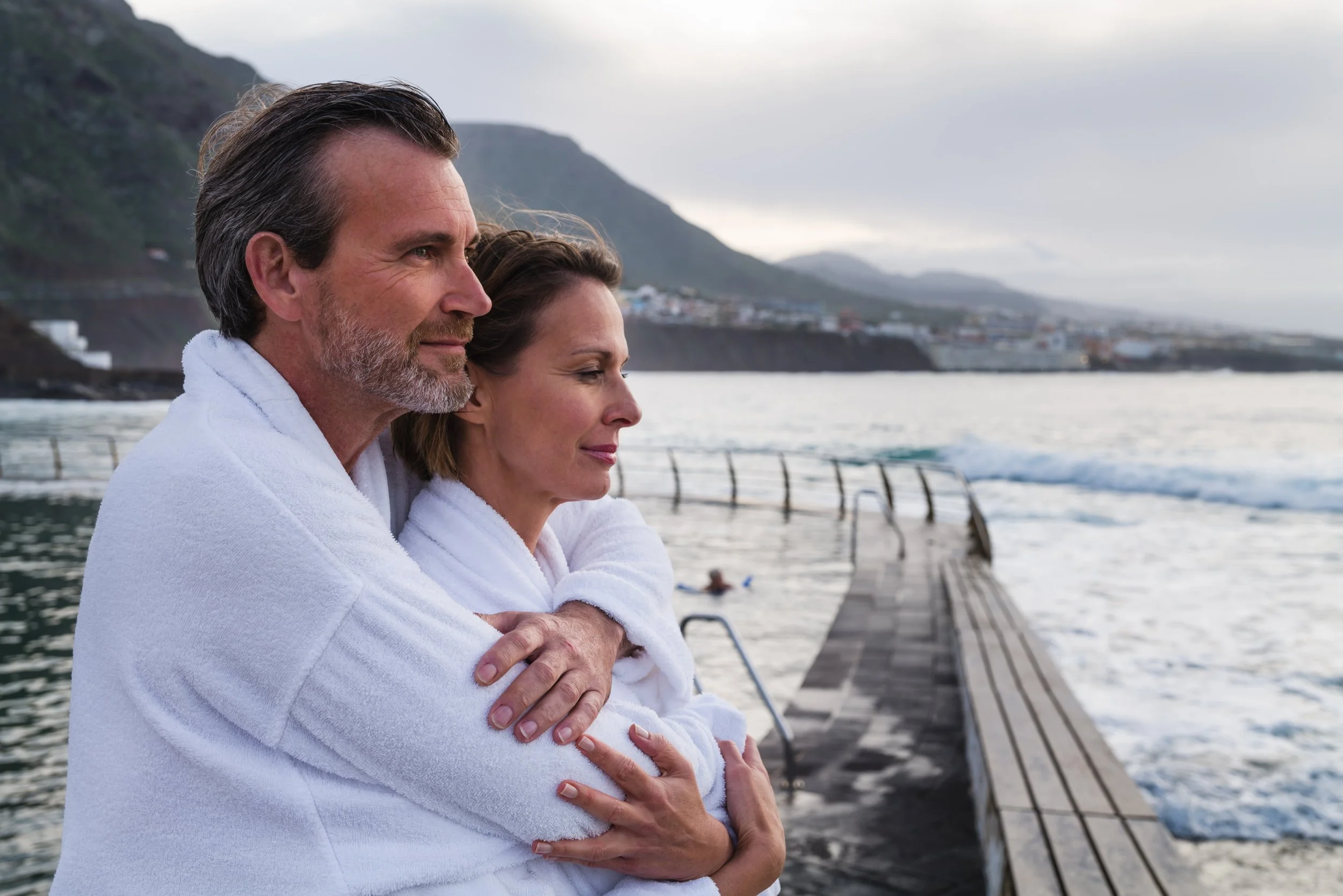 Ein älteres Paar in weißen Bademänteln steht an einer Holzsteg am Meer, umgeben von Bergen, und blickt nach vorn, während die Sonne im Himmel untergeht.