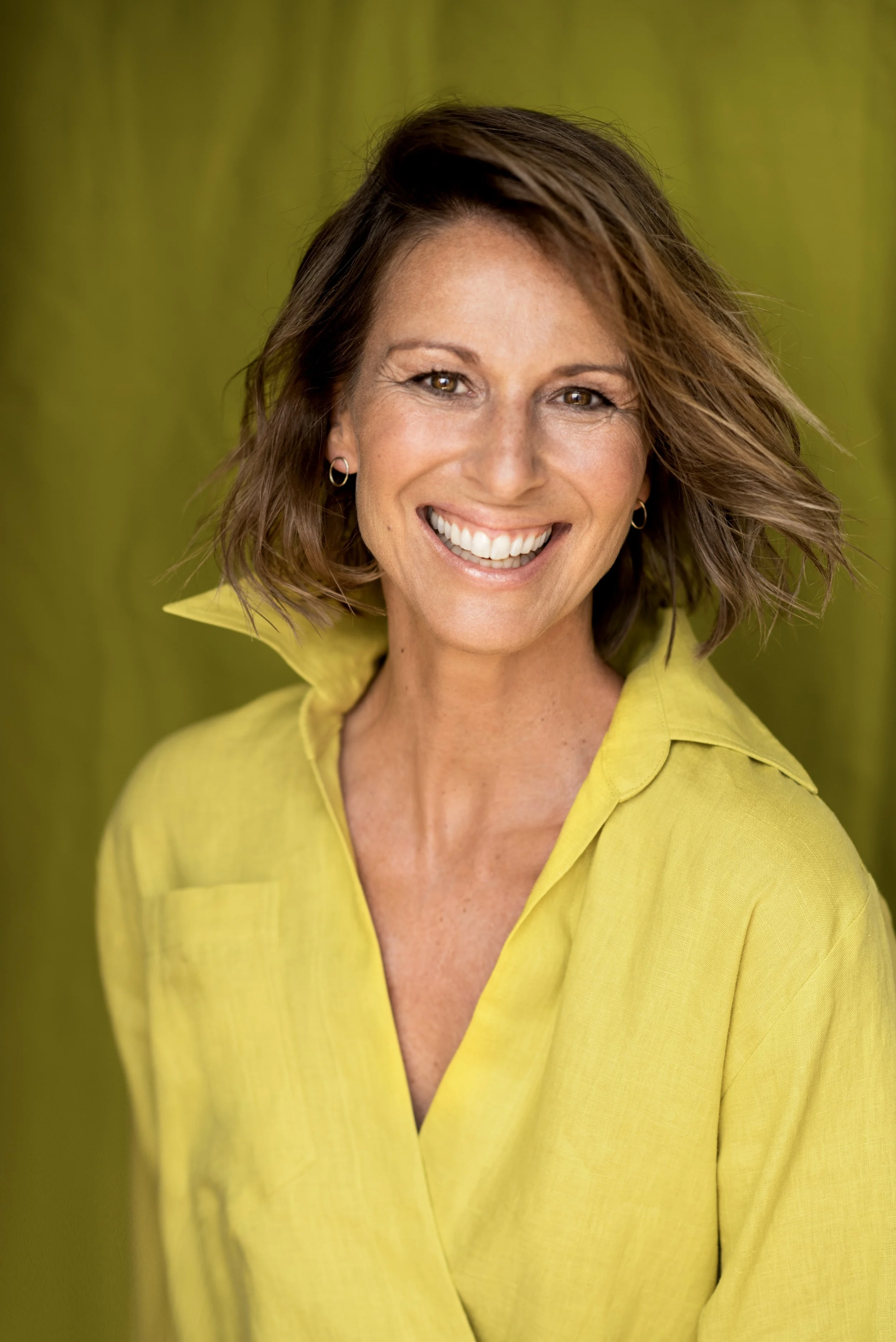 Mittlere Frau mit braunen, lockigen Haaren und einem Lächeln, trägt eine gelbe Bluse vor grünem Hintergrund.