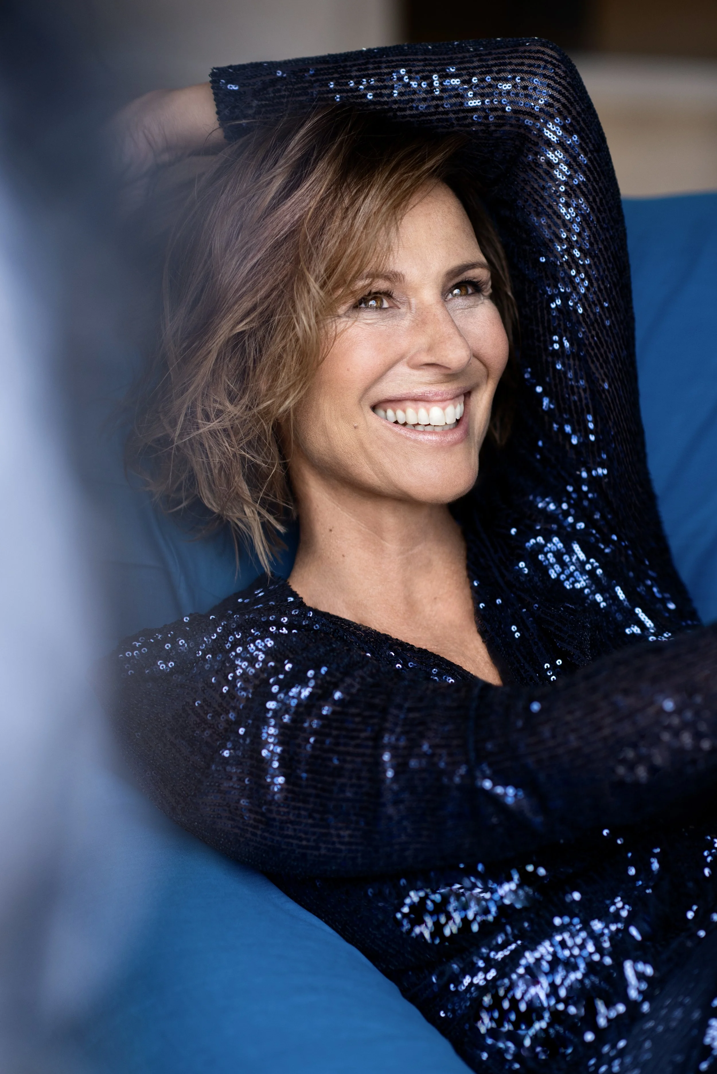 Eine lachende Frau mit braunen, lockigen Haaren, die ein dunkles, mit Sequins besetztes langärmeliges Oberteil trägt, liegt entspannt auf einer blauen Couch.