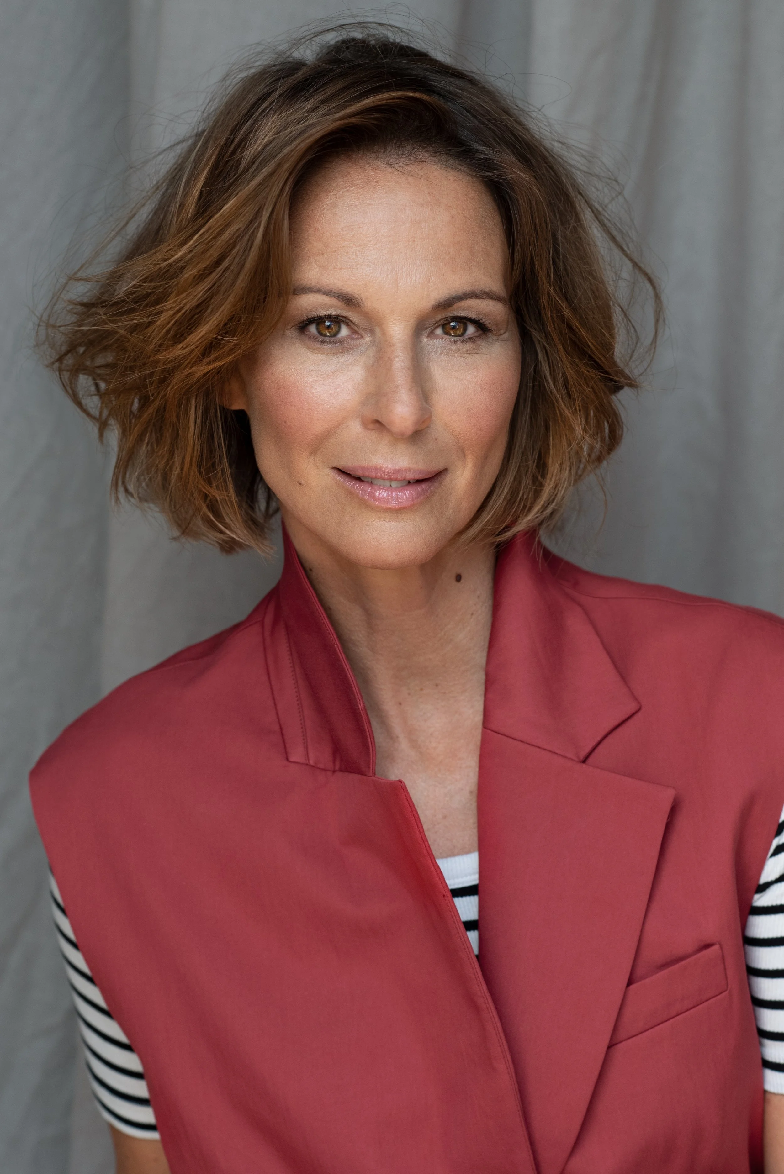 Nahaufnahme einer Frau mit braunen, gewellten Haaren, trägt eine rote Jacke und ein schwarz-weiß gestreiftes Oberteil vor einem grauen Hintergrund.