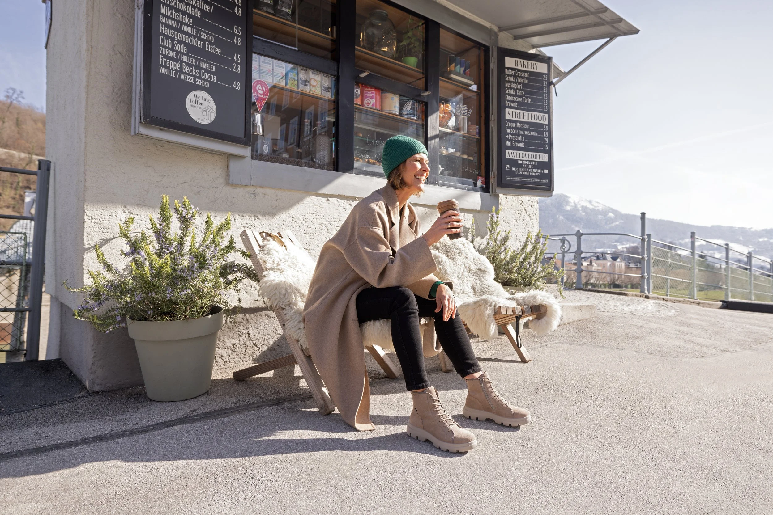 Eine Frau sitzt im Freien vor einem Café, trägt einen grünen Hut, beige Mantel, schwarze Hose und robuste beige Schuhe, hält einen Kaffeebecher in der Hand. Im Hintergrund sind Berge und ein Himmel mit Wolken zu sehen.