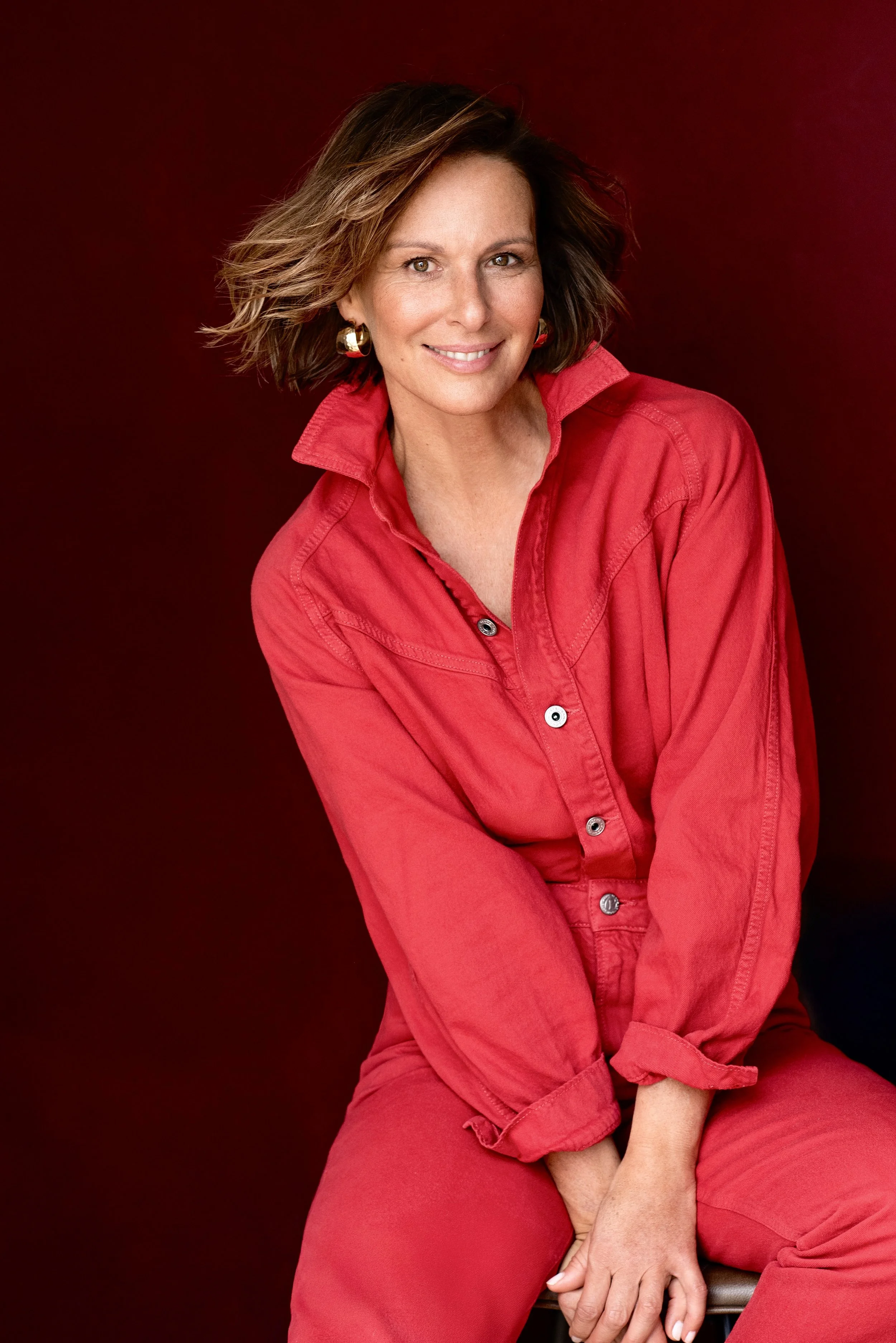 Eine lachende Frau mit kurzen braunen Haaren, die eine rote Bluse und Hose trägt, sitzt vor einem dunklen Hintergrund.