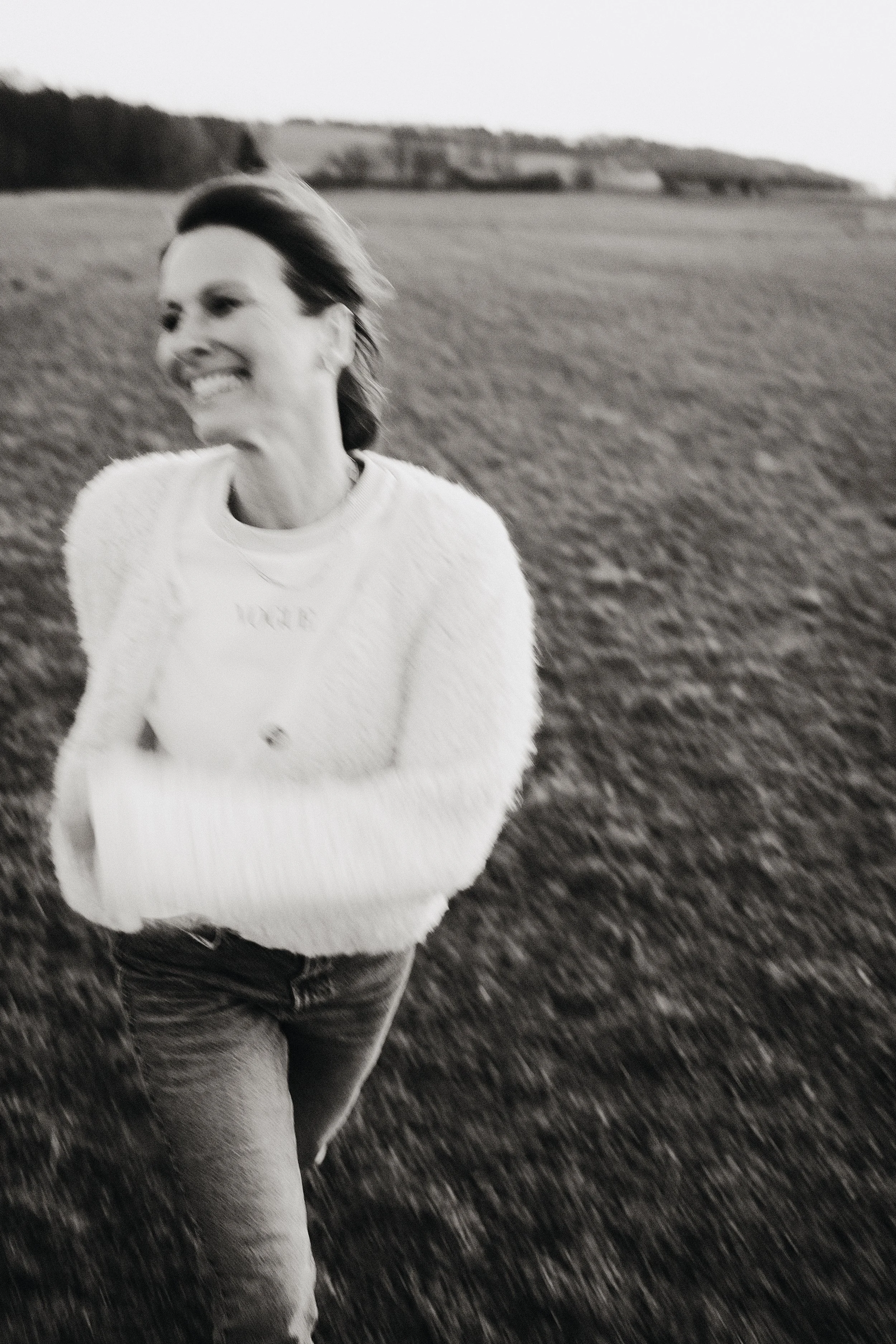 Eine lachende Frau mit dunklen, mittellangen Haaren, die in einem offenen Feld steht, bei Dämmerung, im Schwarz-Weiß-Foto.