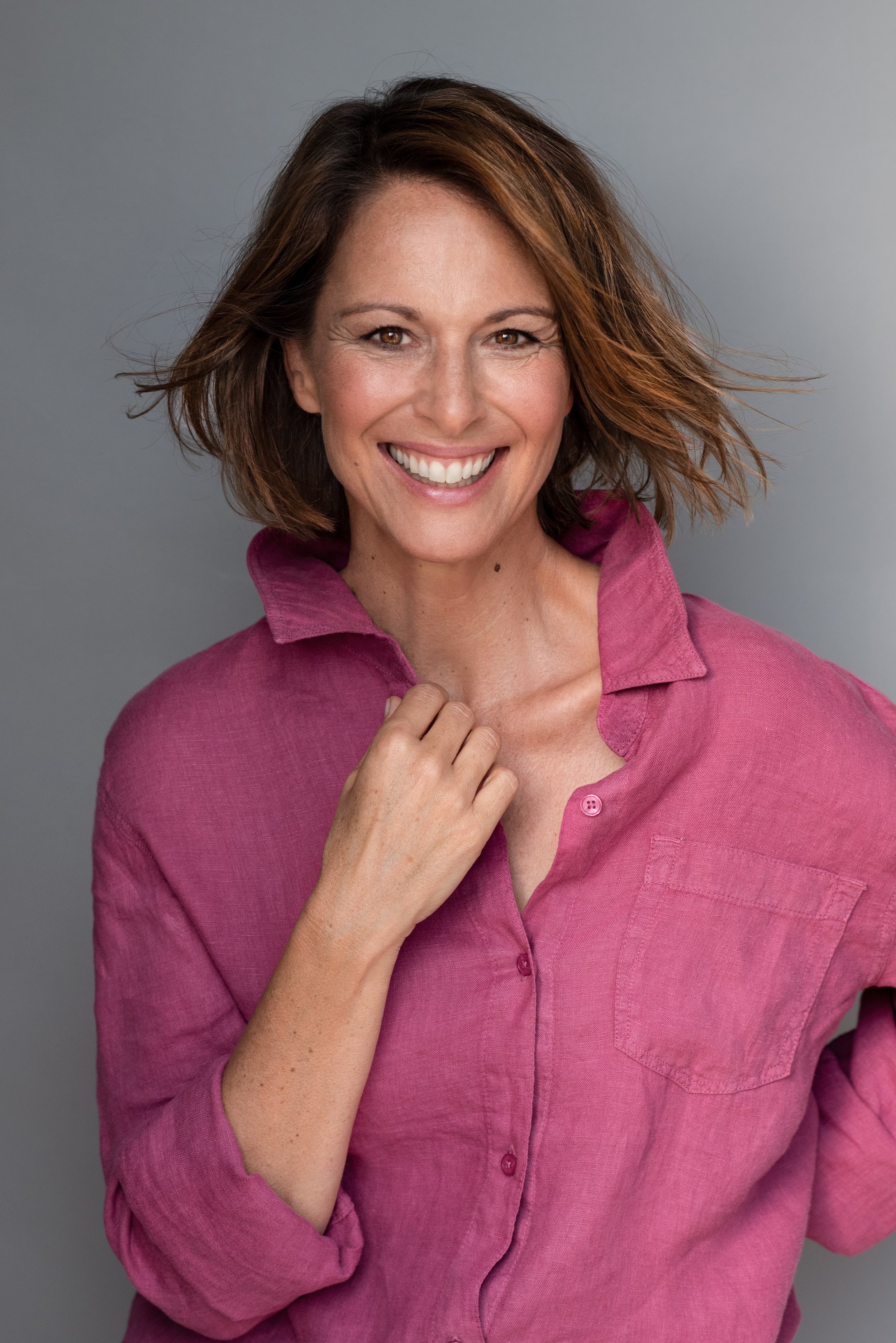 Eine lächelnde Frau mit mittellangen, braunen Haaren trägt eine pinkfarbene, locker sitzende Bluse gegen einen grauen Hintergrund.