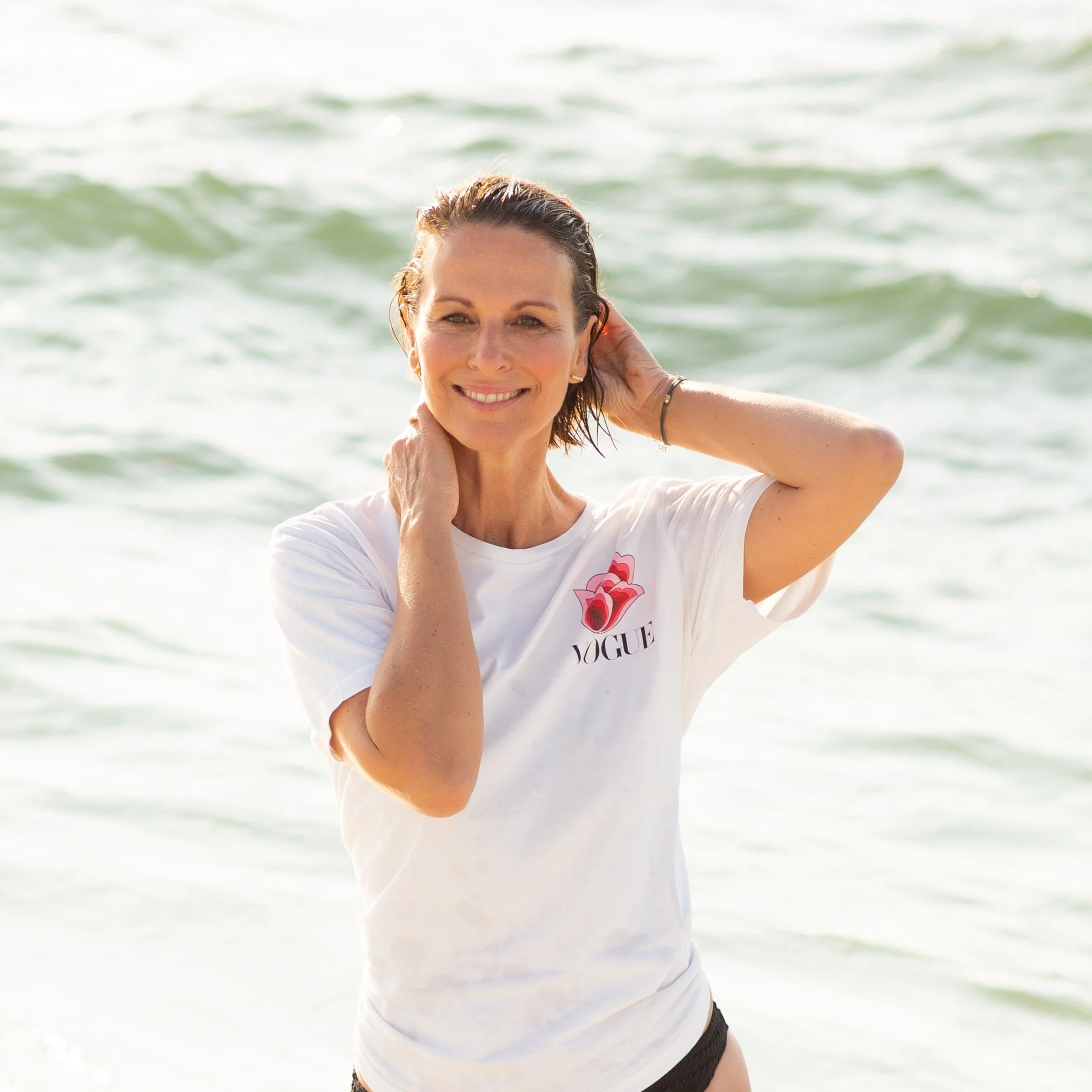 Lächelnde Frau am Strand, trägt ein weißes T-Shirt mit einer Rose und dem Wort Vogue, Wasser im Hintergrund.