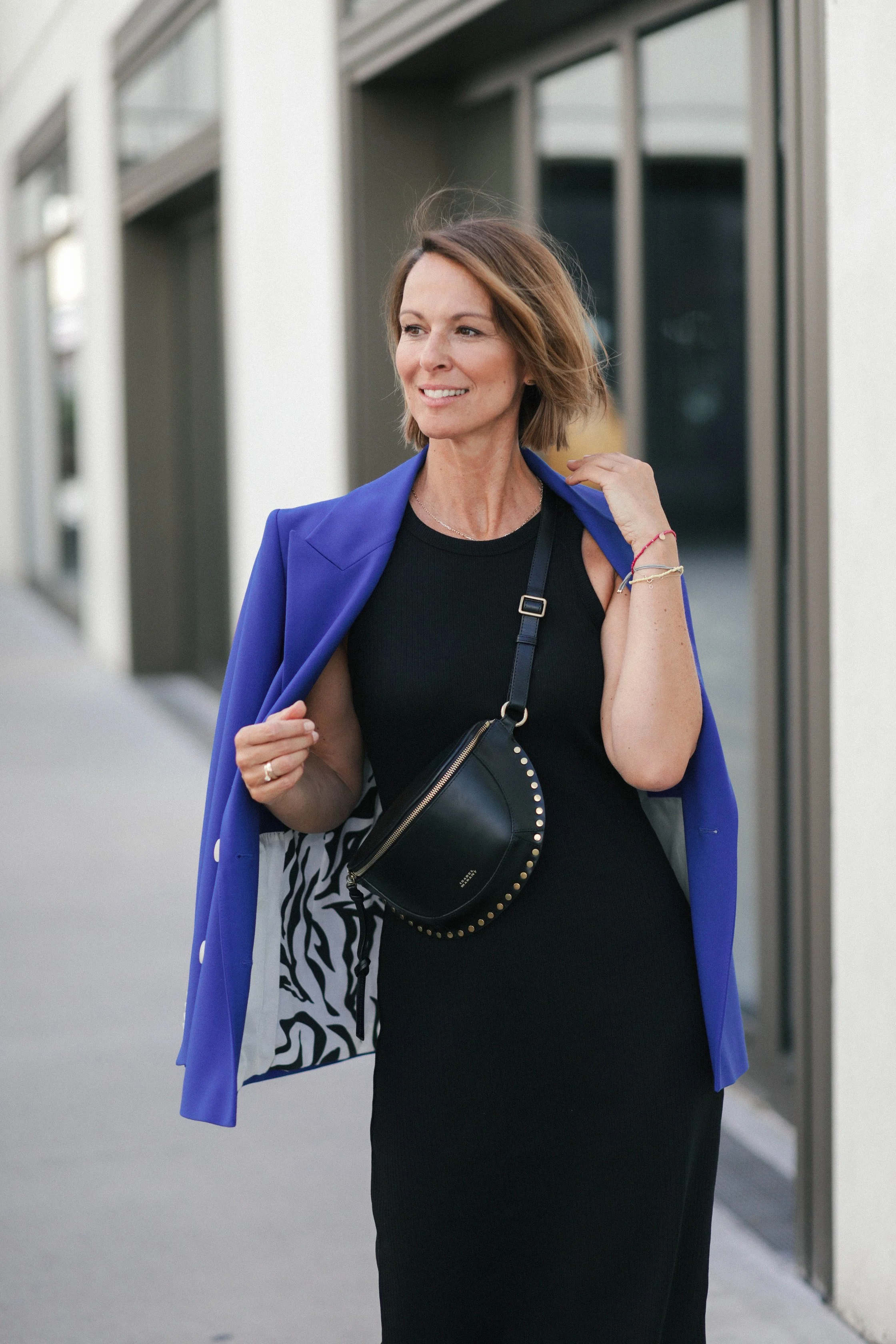 Eine lächelnde Frau in einem schwarzen Kleid mit einem blauen Blazer, die eine kleine schwarze Tasche trägt, während sie an einem modernen Gebäude entlanggeht.