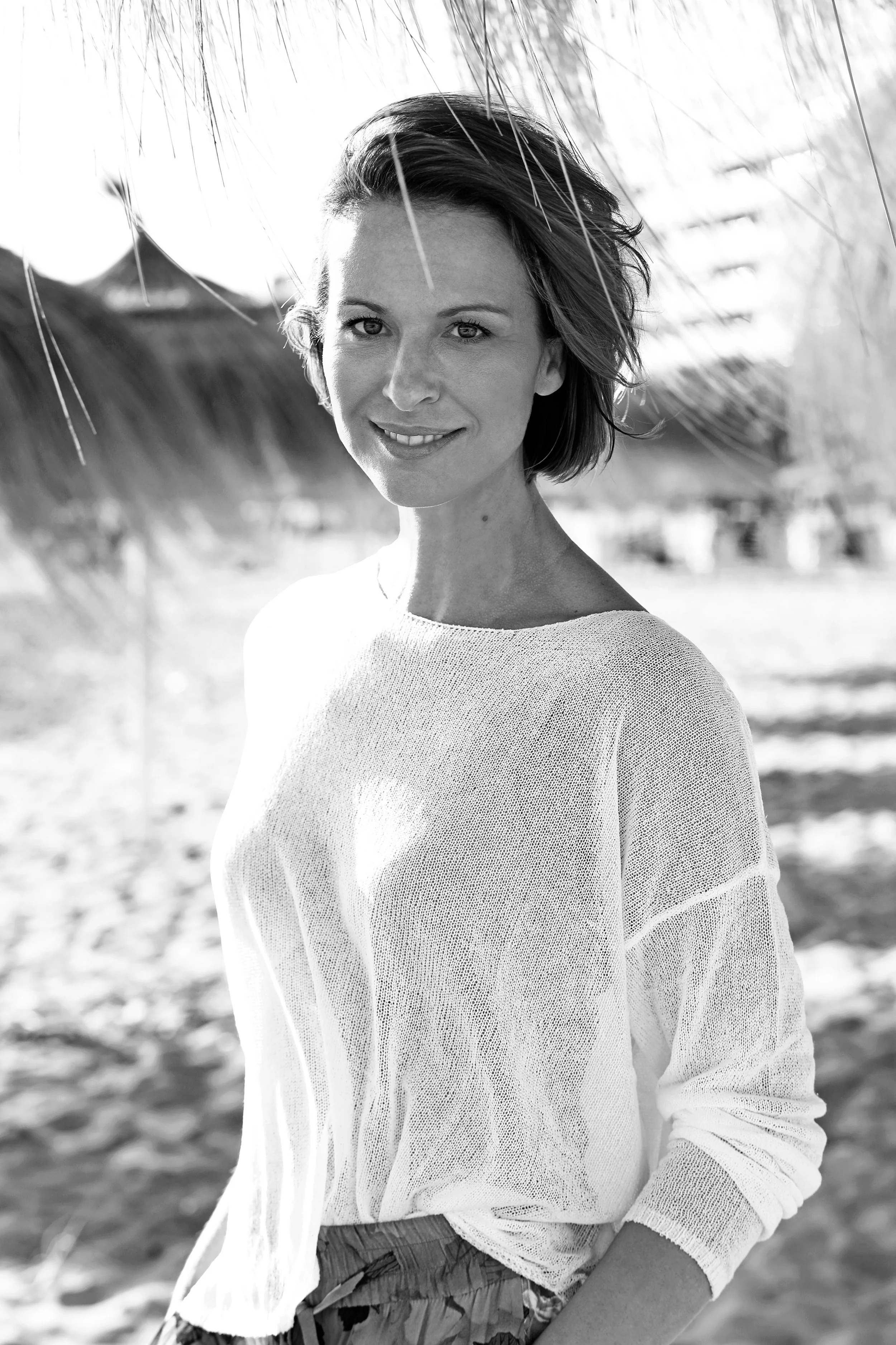 Schwarz-weiß-Foto einer lächelnden Frau mit kurzen Haaren, die an einem Strand unter Palmen steht.