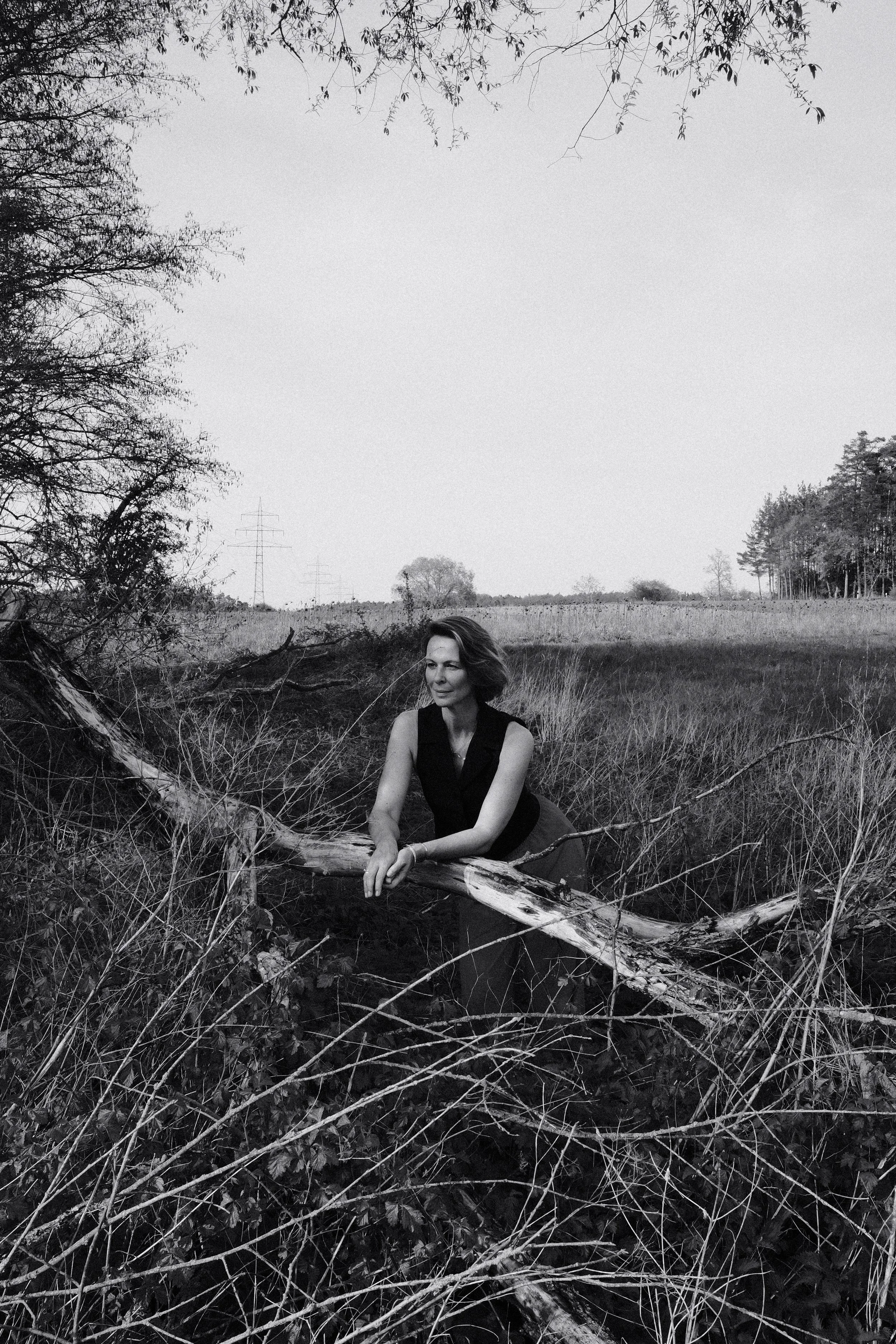 Eine Frau in einem schwarzen Kleid, die auf einem umgestürzten Baum in einer ländlichen, offenen Graslandschaft sitzt, umgeben von vertrockneten Zweigen und Bäumen im Hintergrund, schwarz-weiß fotografiert.