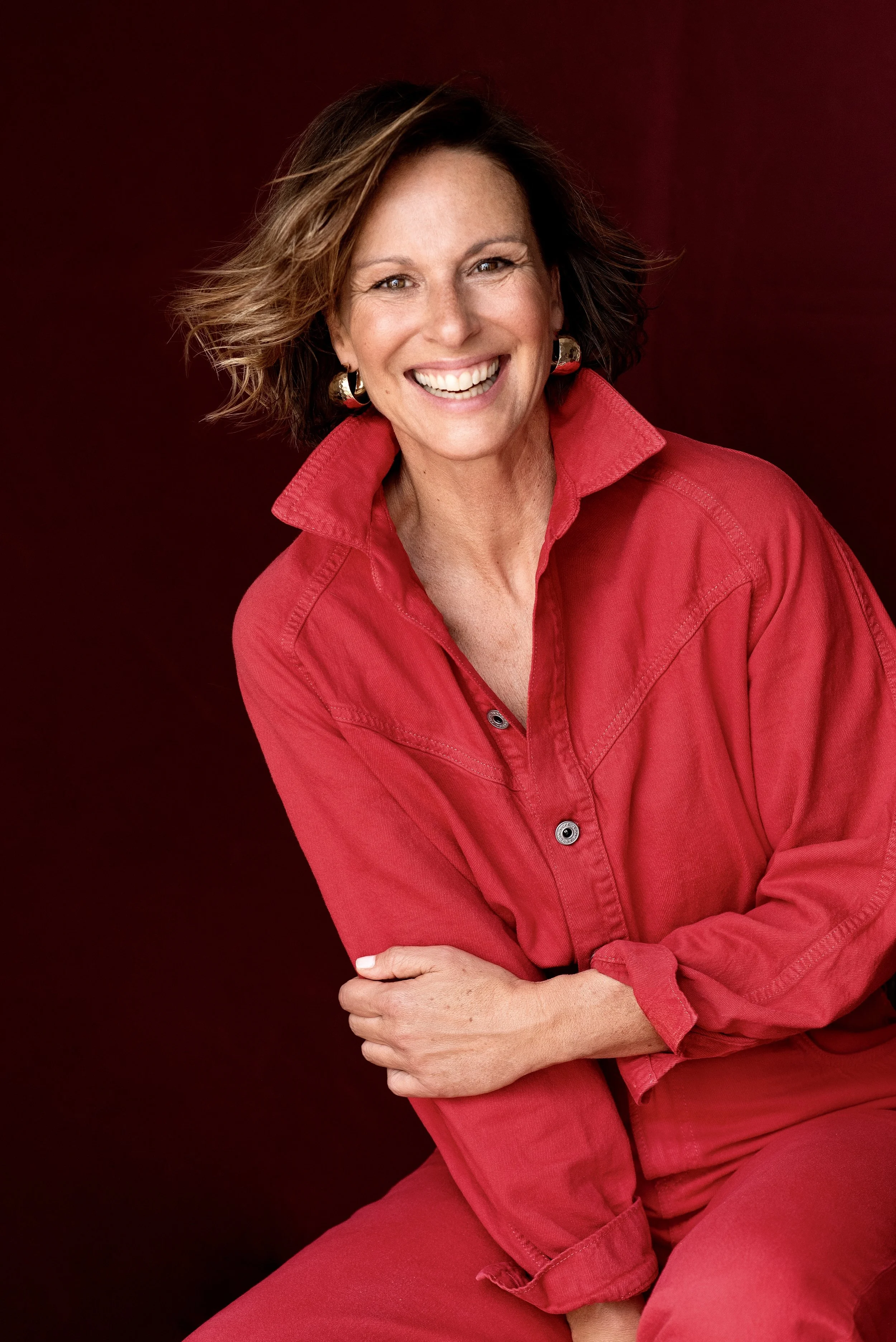 Eine lächelnde Frau mit kurzen braunen Haaren, die einen roten Hemd trägt, gegen einen dunkelroten Hintergrund.