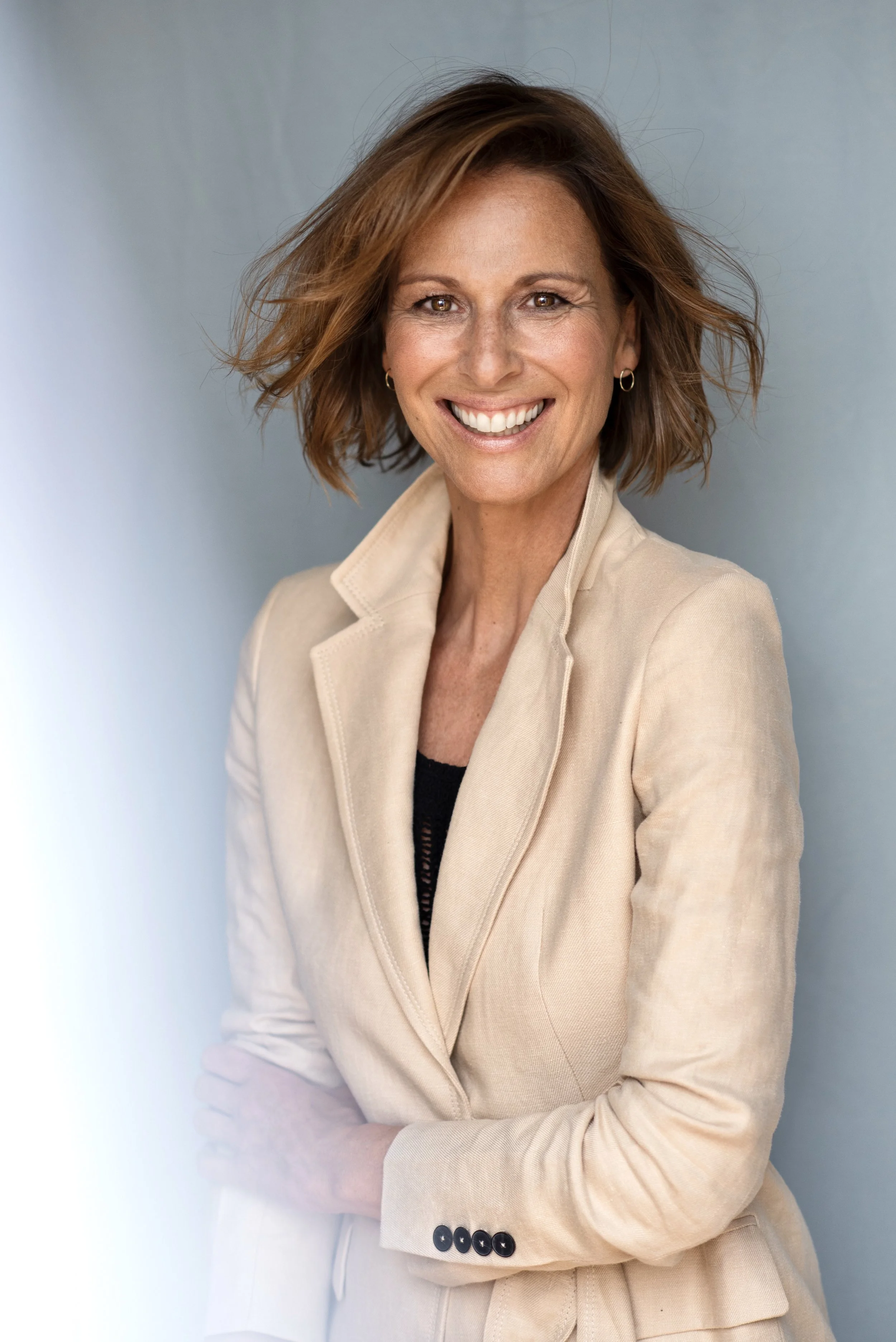 Lächelnde Frau mit kurz lockigem braunem Haar in beige Blazer und schwarzem Top, stehend vor grau Hintergrund.