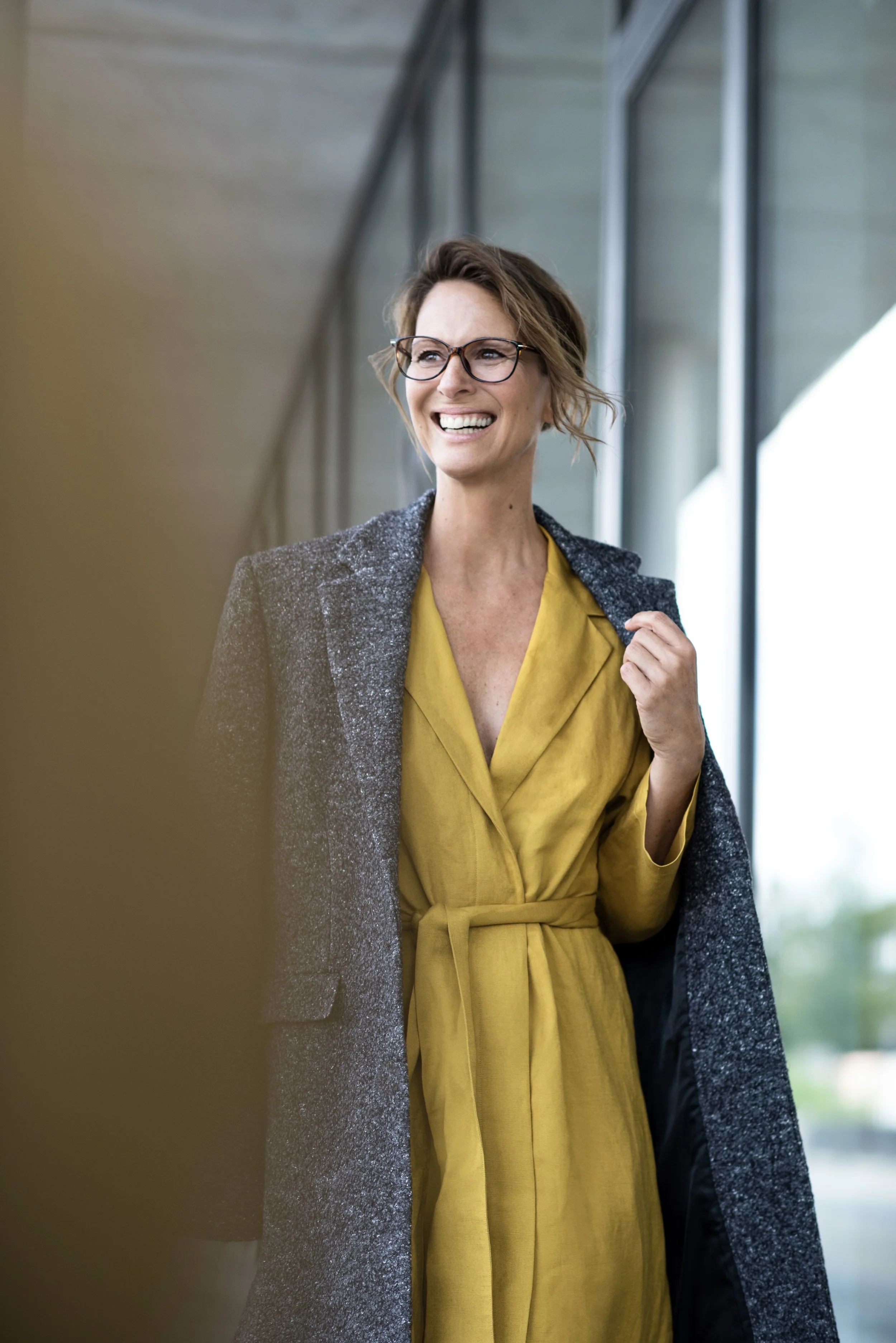Eine lächelnde Frau mit Brille trägt einen gelben Mantel und eine graue Jacke, steht in einem modernen Gebäude.