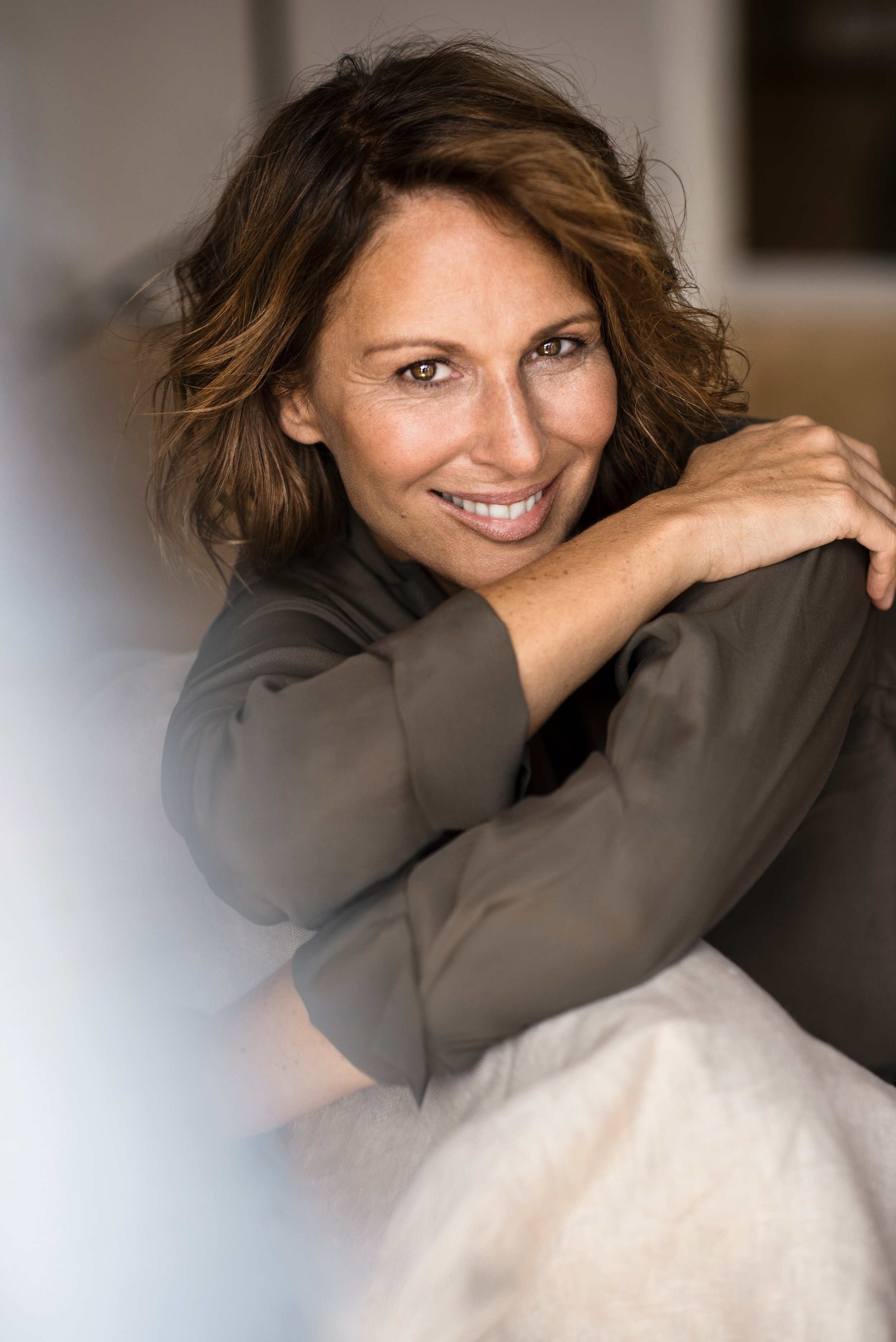 Lächelnde Frau mit braunen, lockigen Haaren, sitzt entspannt auf einem Sofa, trägt eine graue Bluse, im Hintergrund ein Fenster.