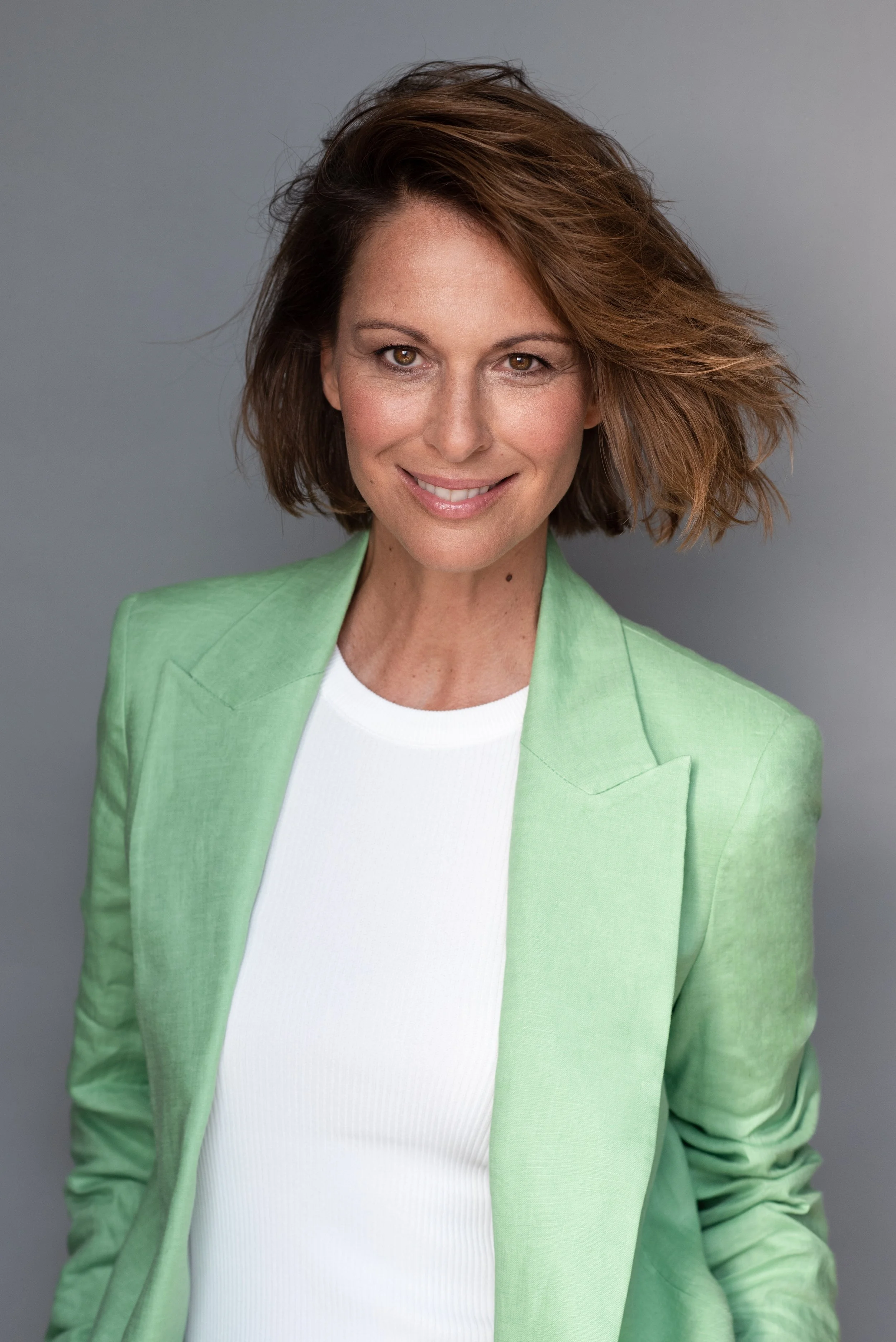 Porträt einer lächelnden Frau mit braunen, lockigen Haaren in einem hellgrünen Blazer vor grauem Hintergrund.