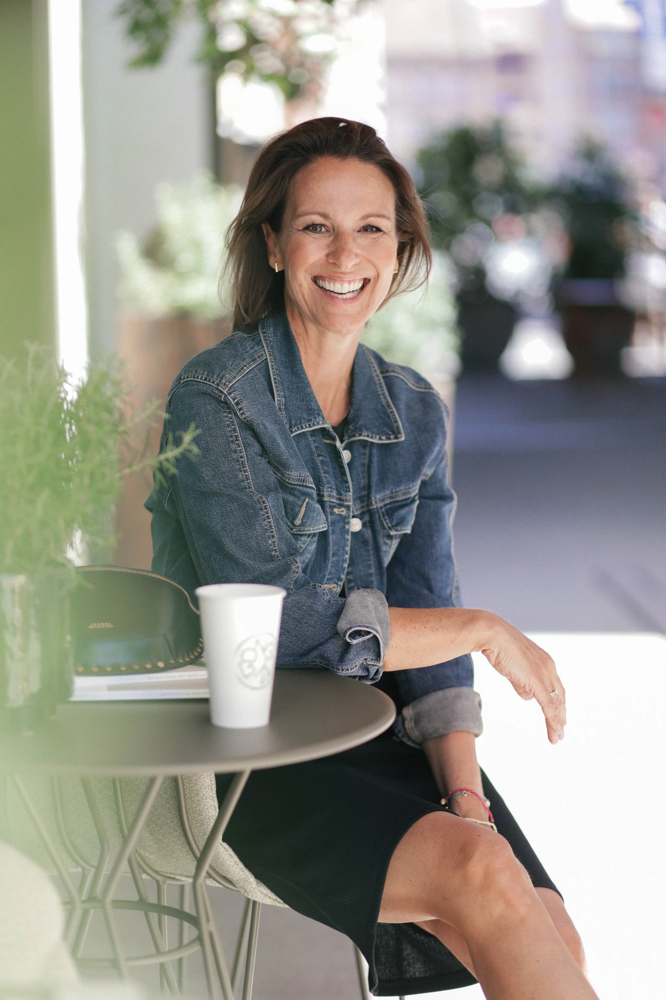 Lächelnde Frau sitzt an einem Tisch im Café, trägt Jeansjacke und schwarzes Kleid, mit Kaffee to-go auf dem Tisch.