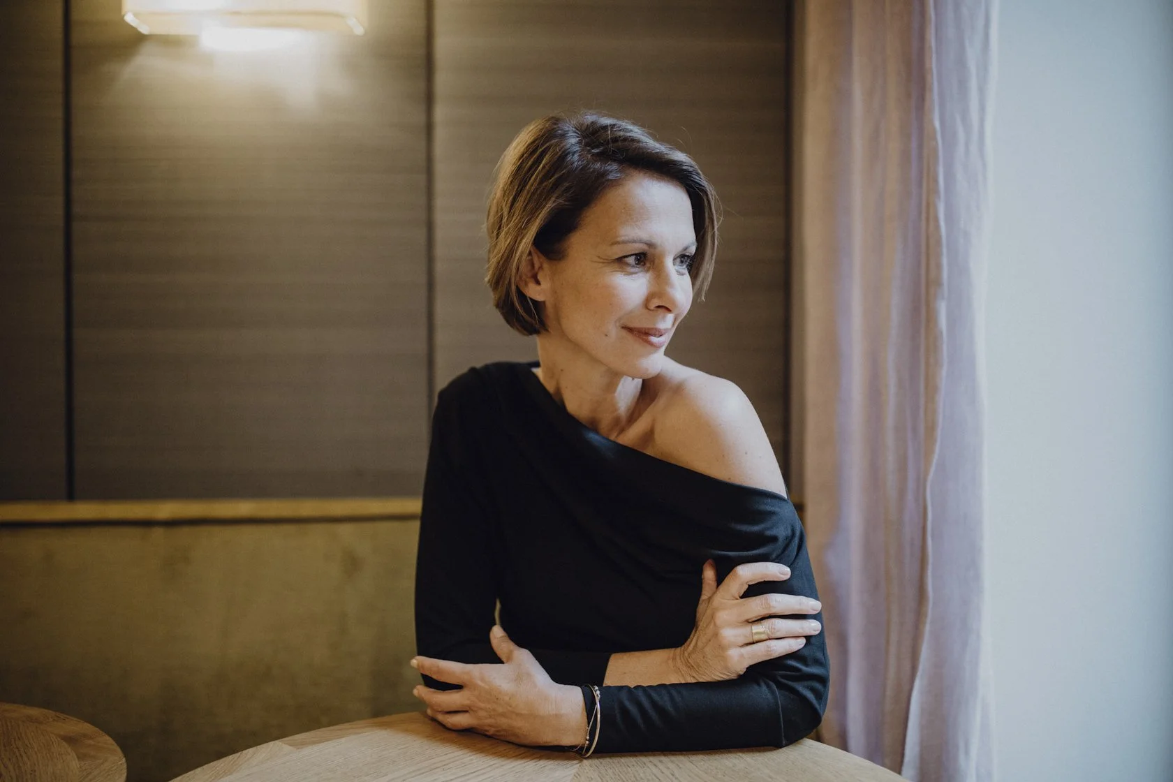 Eine Frau mit kurzen braunen Haaren, die ein schwarzes ein Schulter-Top trägt, sitzt an einem Tisch in einem Raum mit Holzvertäfelung und guckt nach draußen.