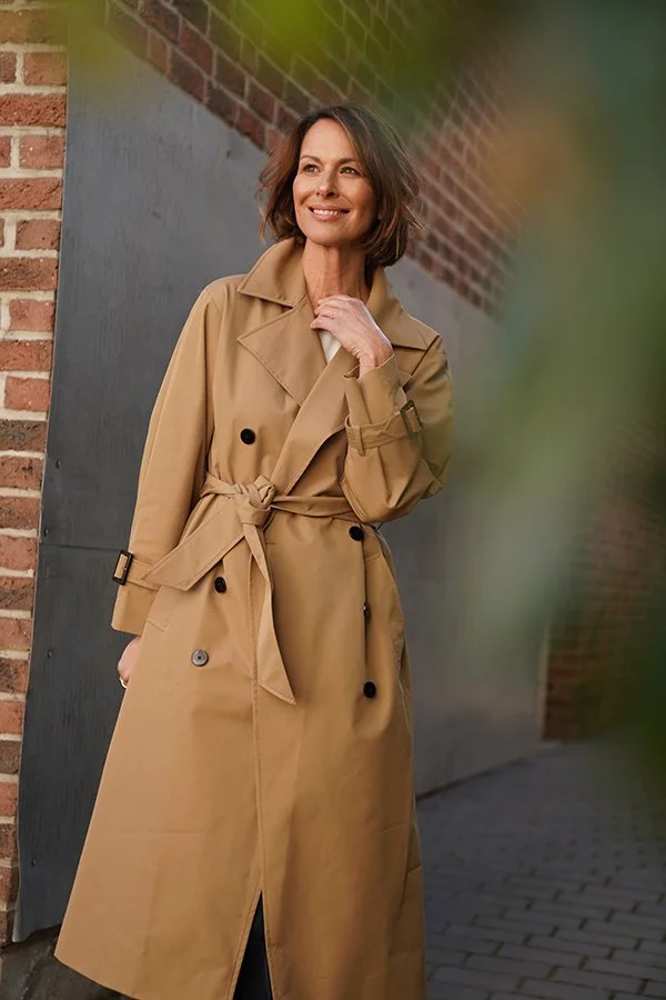 Frau in beigem Trenchcoat steht an einer Mauer, lächelt, Blick nach oben, im Hintergrund Ziegelwand.