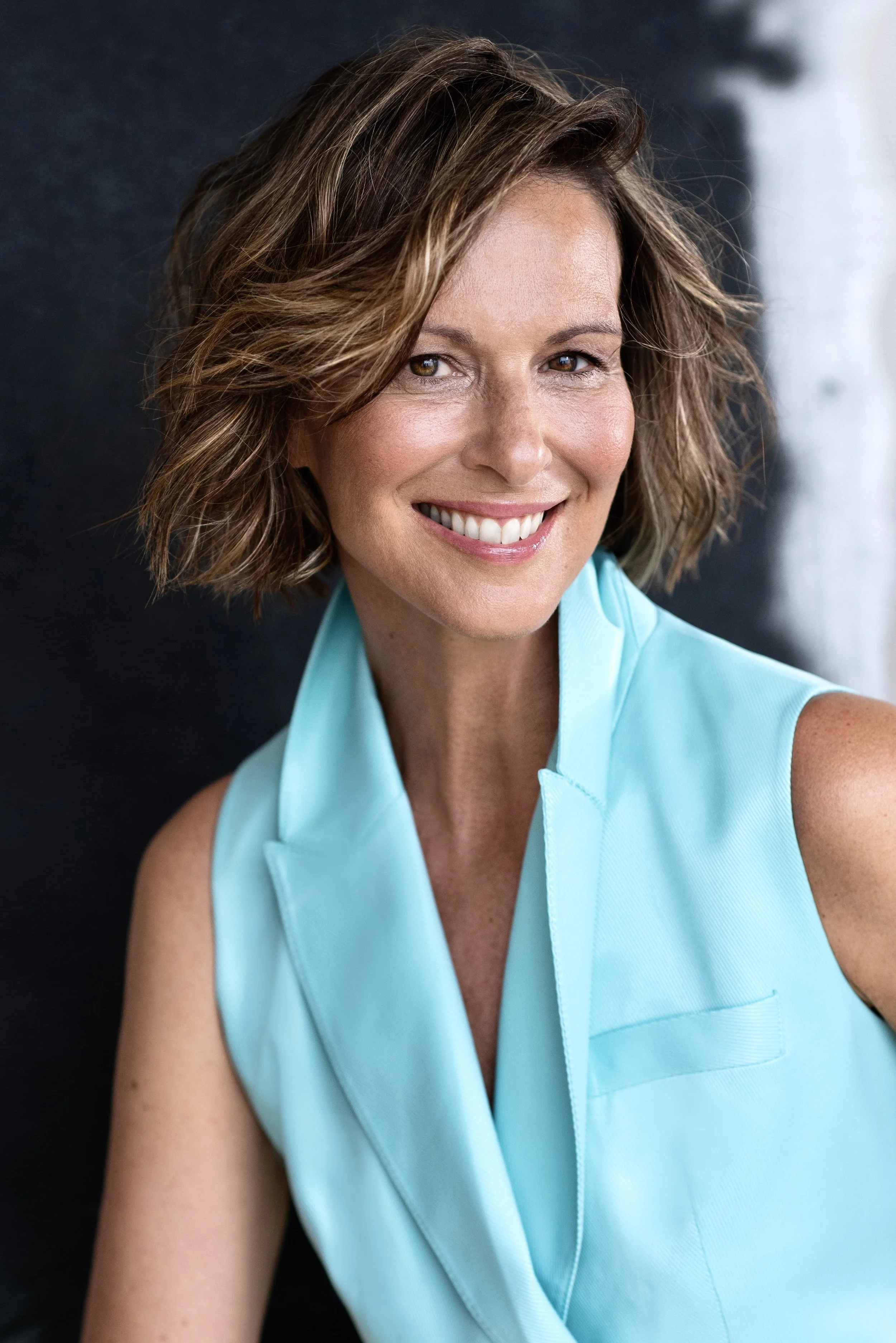 Lächelnde Frau mit braunen, gewellten Haaren in einem hellblauen ärmellosen Top vor dunklem Hintergrund.