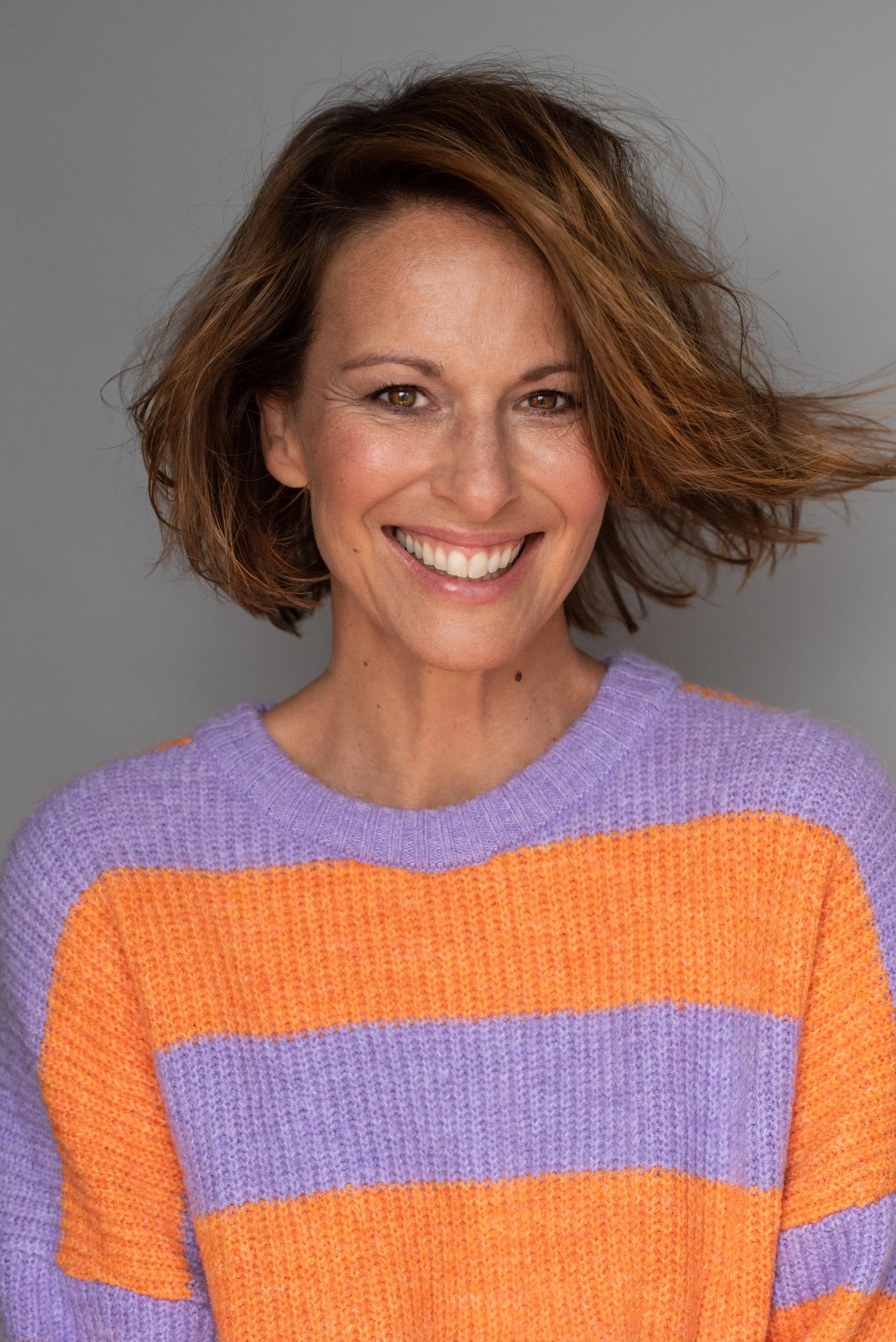 Lächelnde Frau mit kurzen braunen Haaren in lila und orange gestreiftem Pullover gegen grauen Hintergrund.