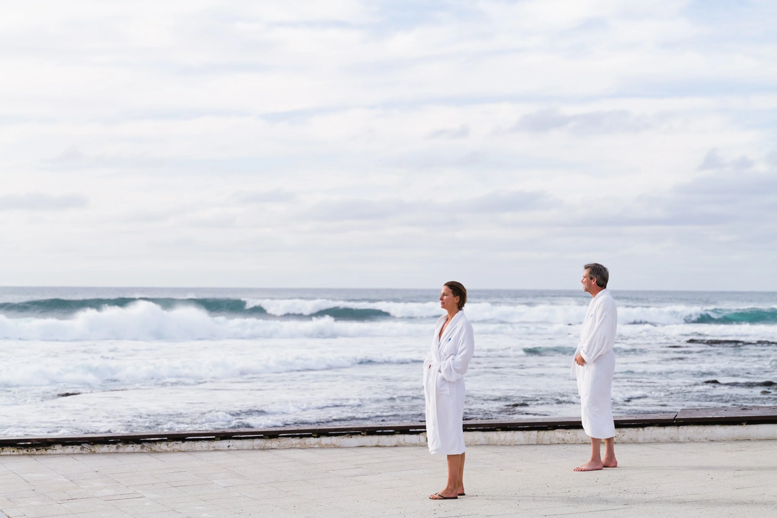 Zwei Personen im Bademantel stehen am Strand, schauen aufs Meer, Wolken am Himmel.