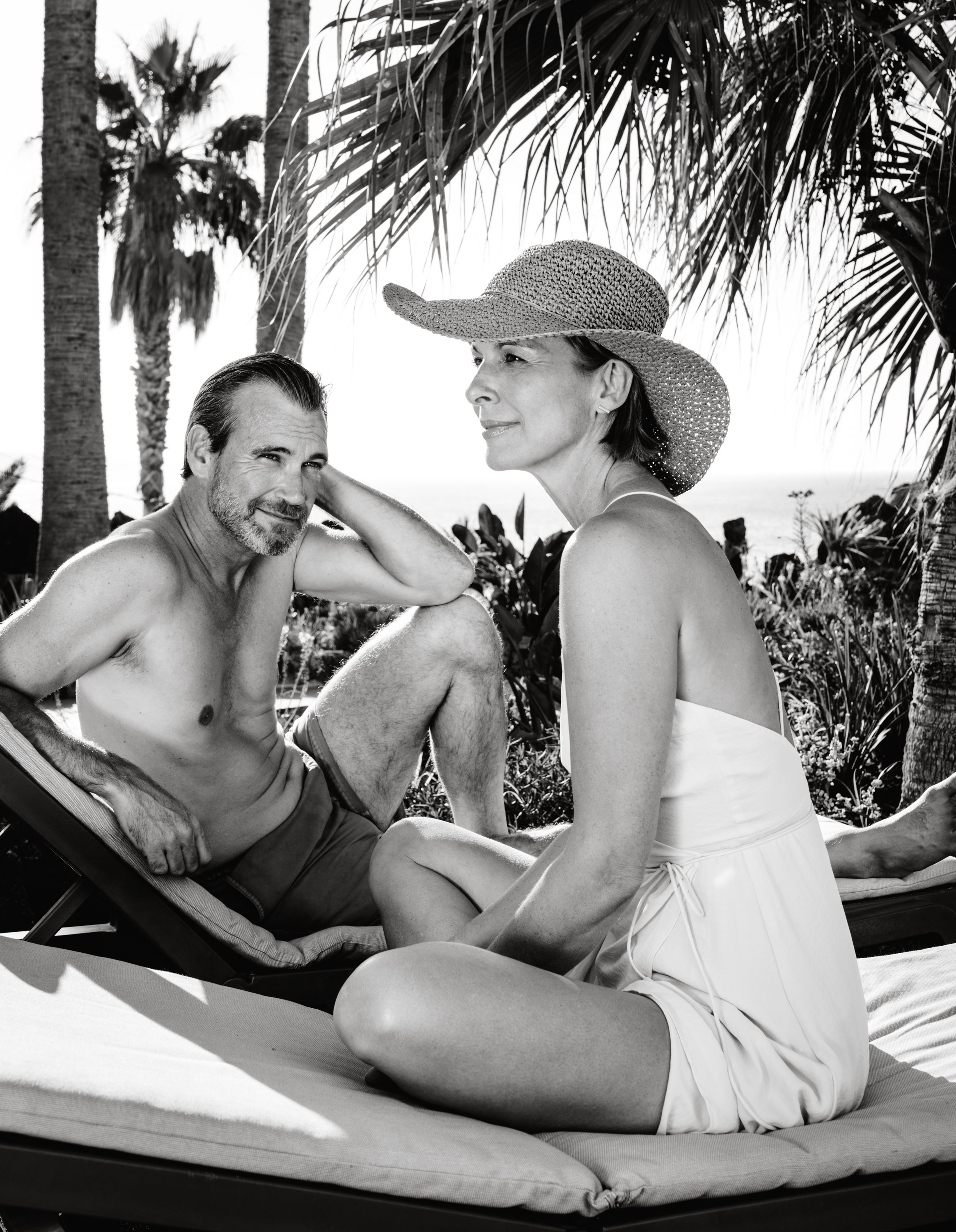 Ein Mann und eine Frau entspannen sich im Freien auf einem Liegestuhl, umgeben von Palmen, bei sonnigem Wetter.