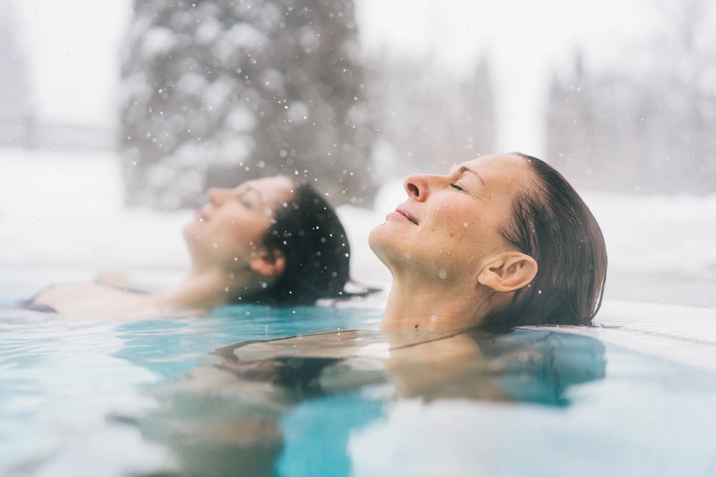 Zwei Frauen entspannen im Pool mit geschlossenen Augen, während es draußen schneit.