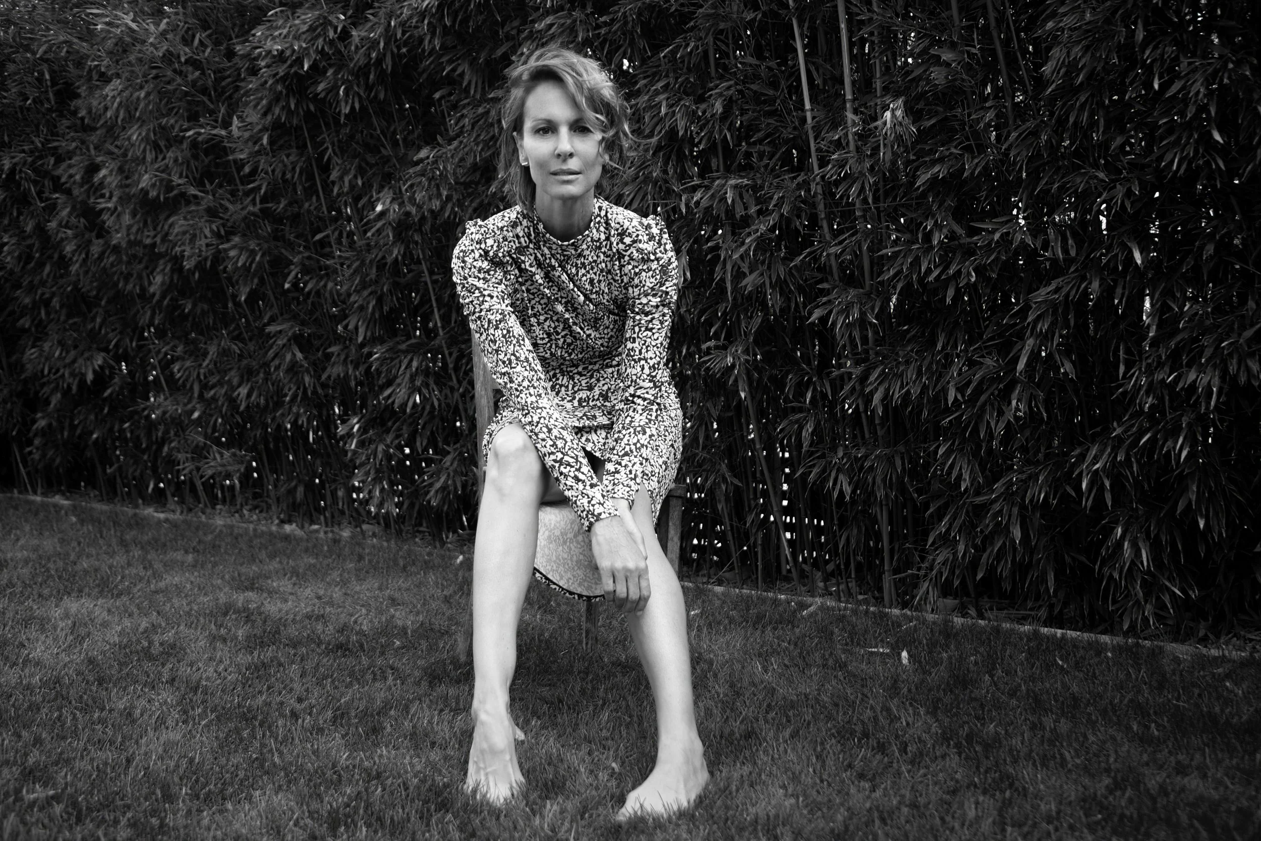 Schwarz-weiß-Fotografie einer Frau, die barfuß auf einem Rasen sitzt, vor einem dichten Busch. Sie trägt ein langärmliges, gemustertes Kleid und schaut direkt in die Kamera.