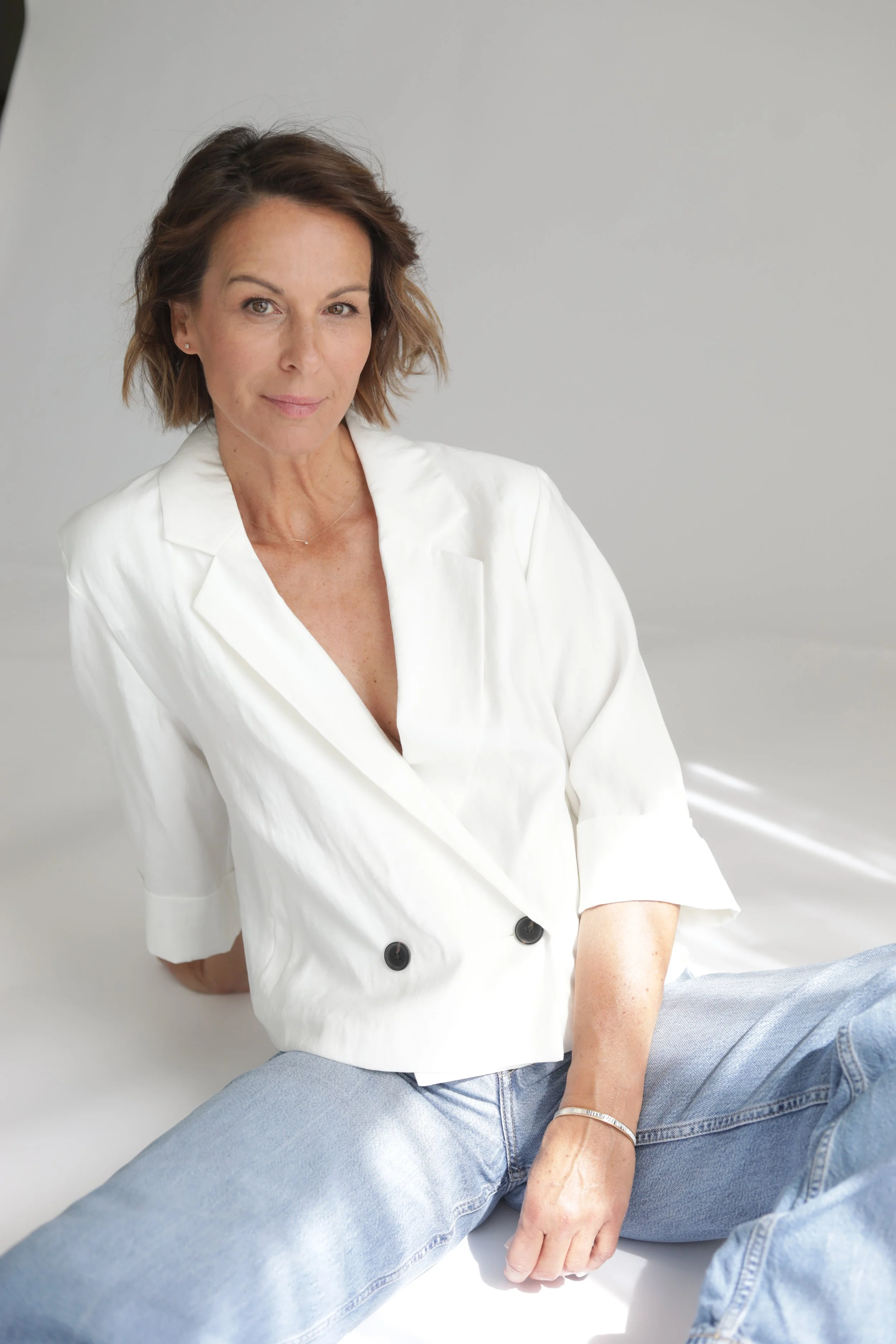 Eine Frau mit kurzen braunen Haaren, die lächelnd sitzend in einem weißen Blazer und blauen Jeans vor einem neutralen Hintergrund sitzt.