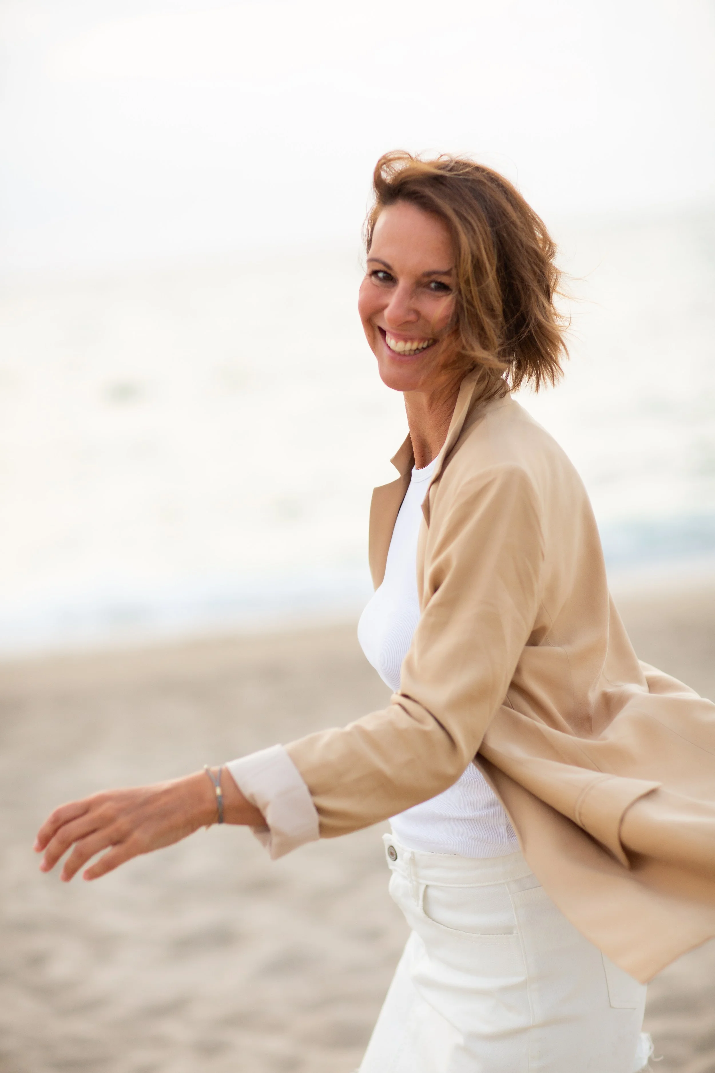 Eine lachende Frau mit kurzen braunen Haaren am Strand bei Sonnenuntergang.