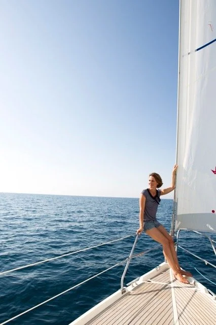 Junge Frau sitzt auf dem Bug eines Segelbootes im Meer, hält das Segel fest, bei sonnigem Himmel.