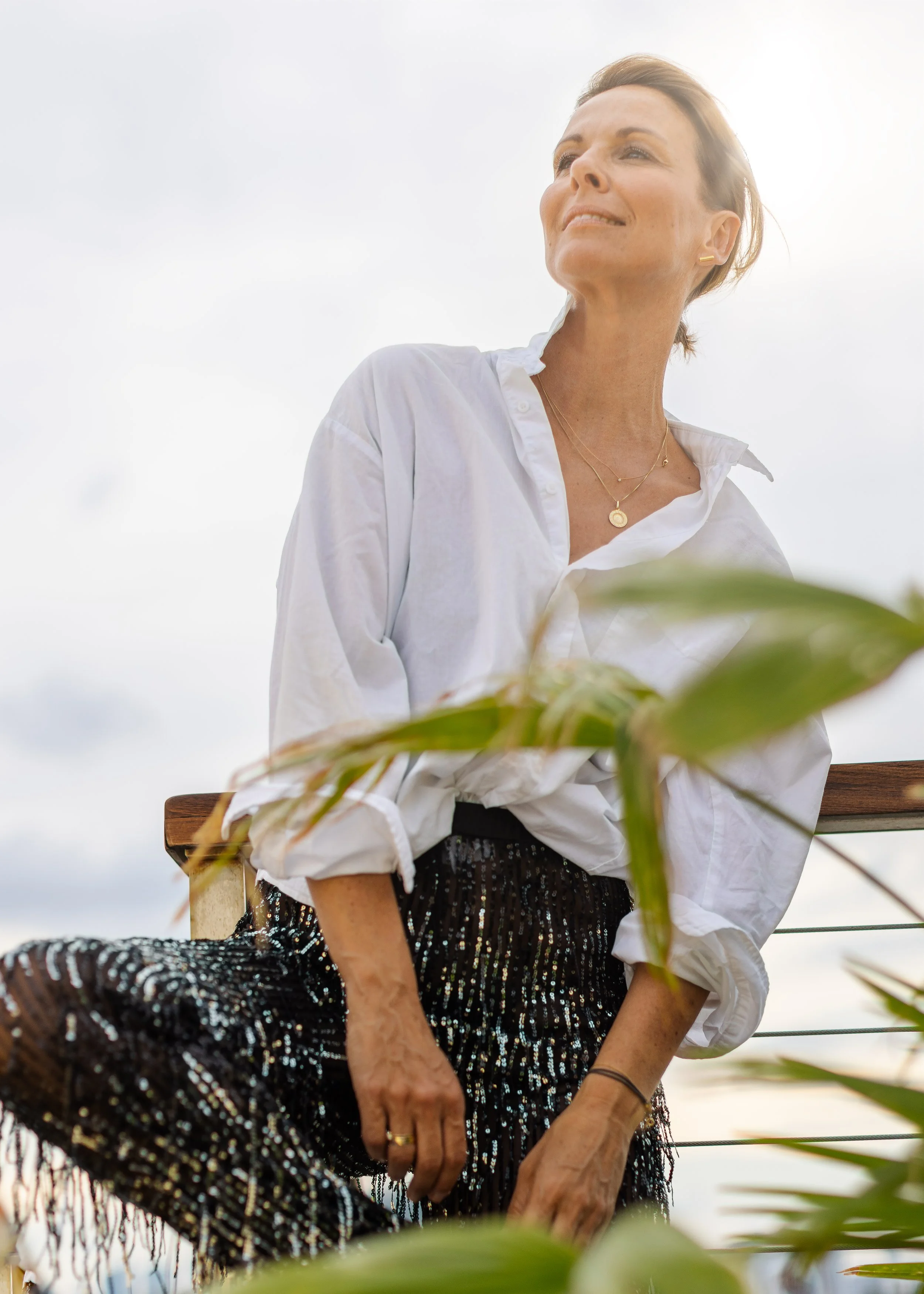 Eine Frau in einer weißen Bluse und einem schwarzen, glitzernden Rock steht auf einem Balkon im Freien, blickt nach oben und lächelt leicht, mit Wolken im Himmel im Hintergrund.