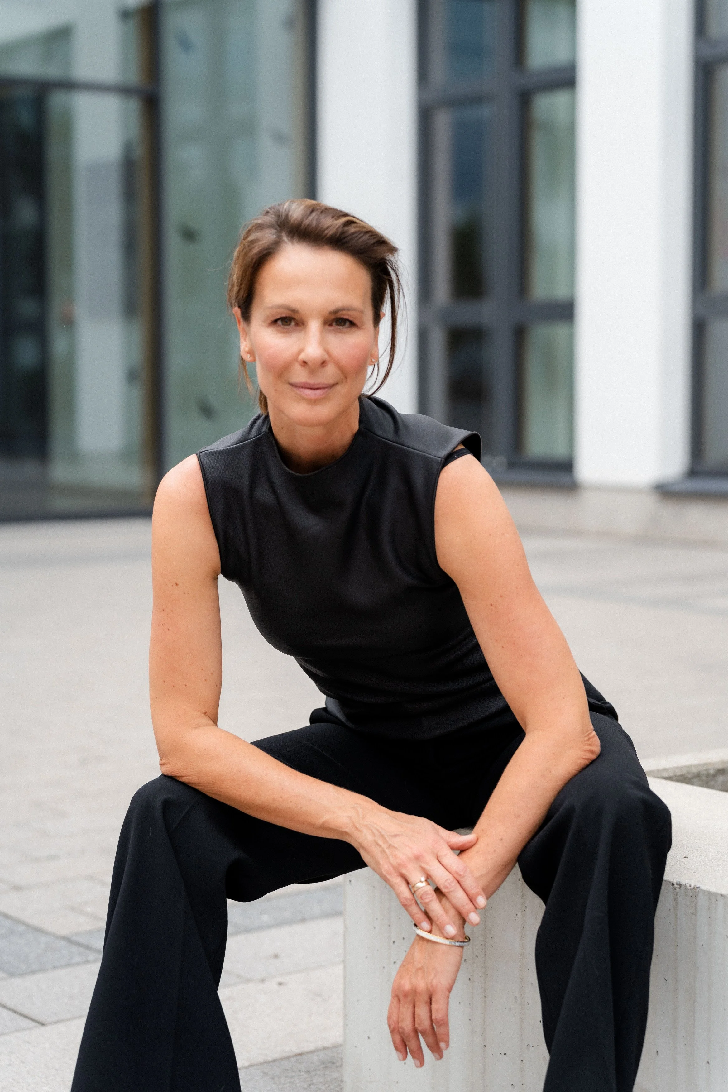 Eine Frau sitzt auf einem Betonblock vor einem modernen Gebäude, trägt eine schwarze ärmellose Bluse und schwarze Hosen, hat kurze braune Haare und schaut in die Kamera.