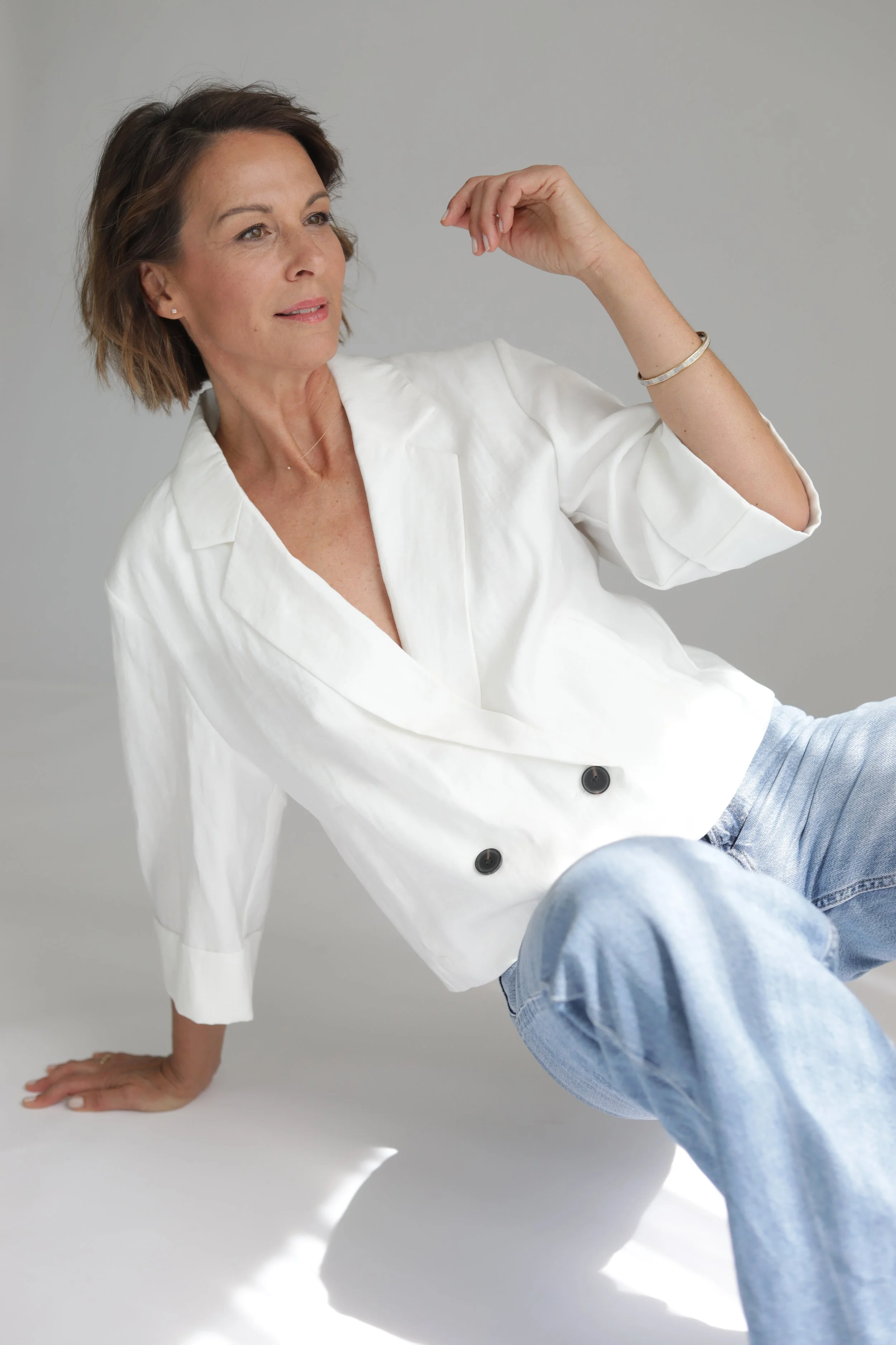 Eine Frau sitzt auf dem Boden, trägt eine weiße Jacke und blaue Jeans, betrachtet nachdenklich, die rechte Hand berührt den Kopf, der Hintergrund ist grau.