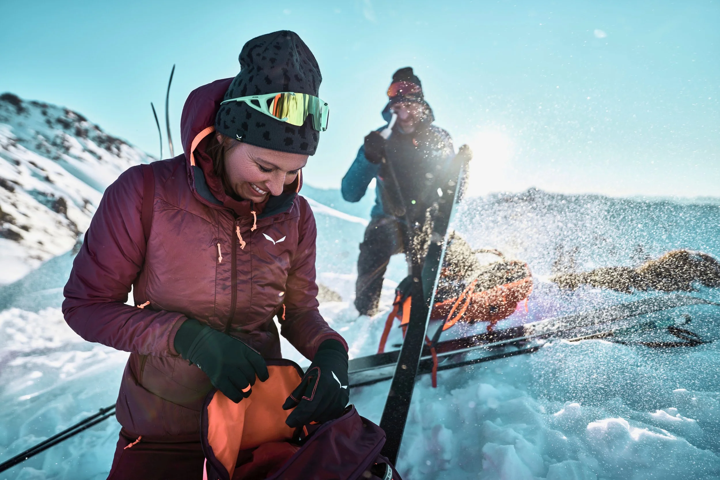 Zwei Menschen in Winterkleidung auf Schneefeld, einer öffnet eine Tasche, während der andere mit einem Schneeschuh auf dem Rücken und Ausrüstung im Schnee steht, bei Sonnenschein.