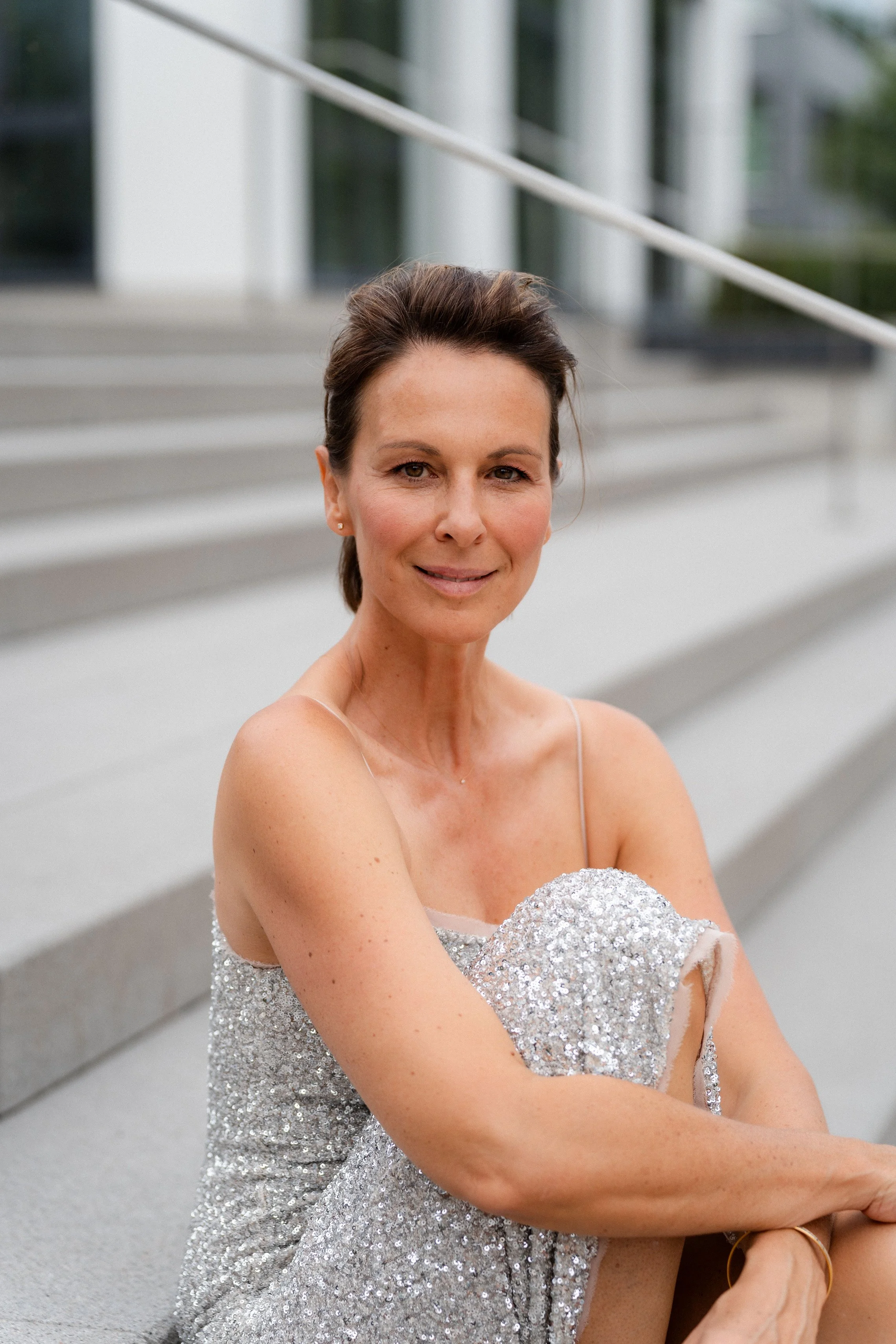 Eine elegant gekleidete Frau sitzt auf den Stufen vor einem modernen Gebäude, trägt ein glitzerndes Kleid und lächelt leicht.