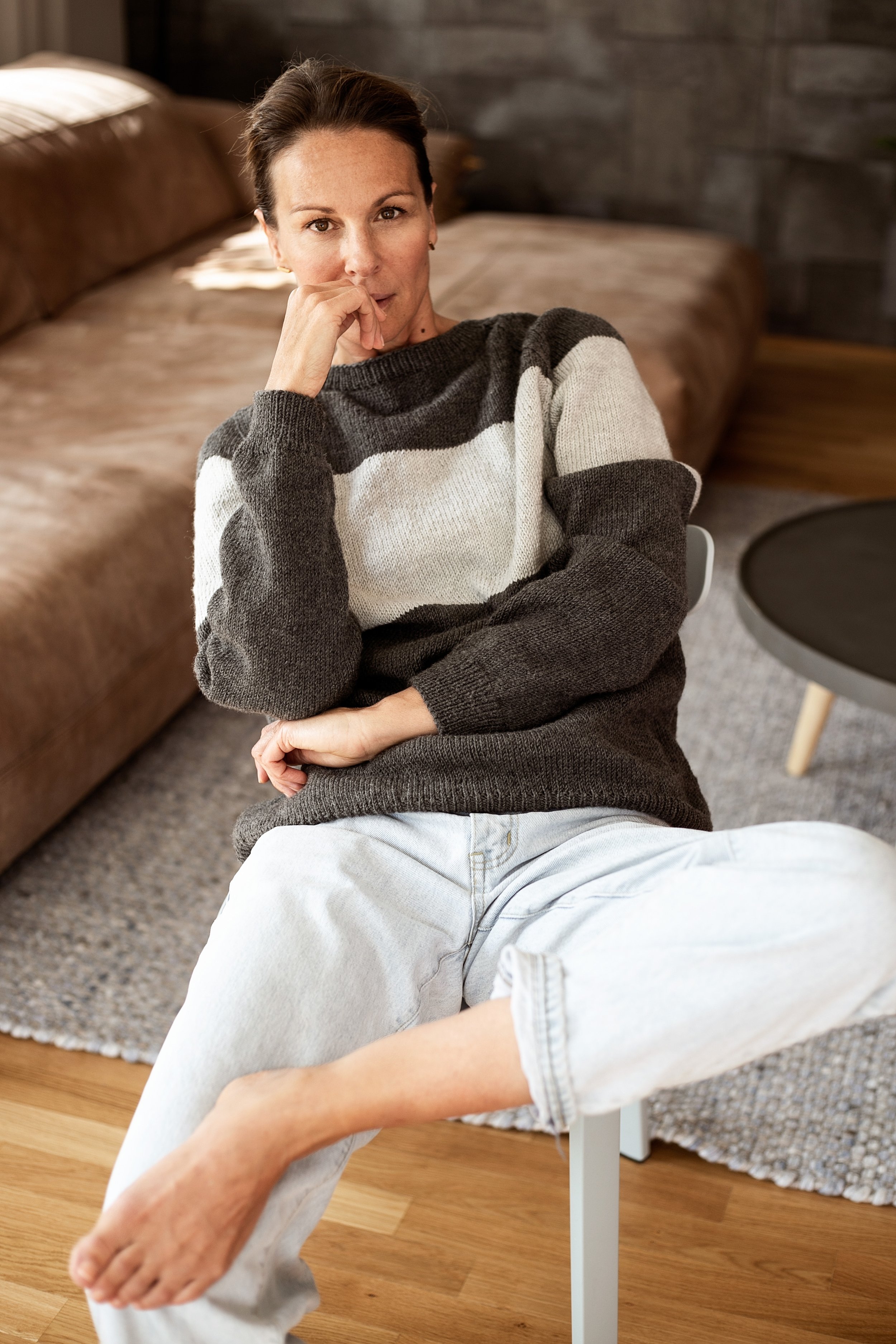 Eine Frau sitzt entspannt auf einem weißen Stuhl in einem Wohnzimmer, trägt einen schwarzen, weißen und grauen Pullover und weiße Jeans, hat braune Haare, betrachtet nachdenklich die Kamera und hat den Ellenbogen auf den Knien abstützt.