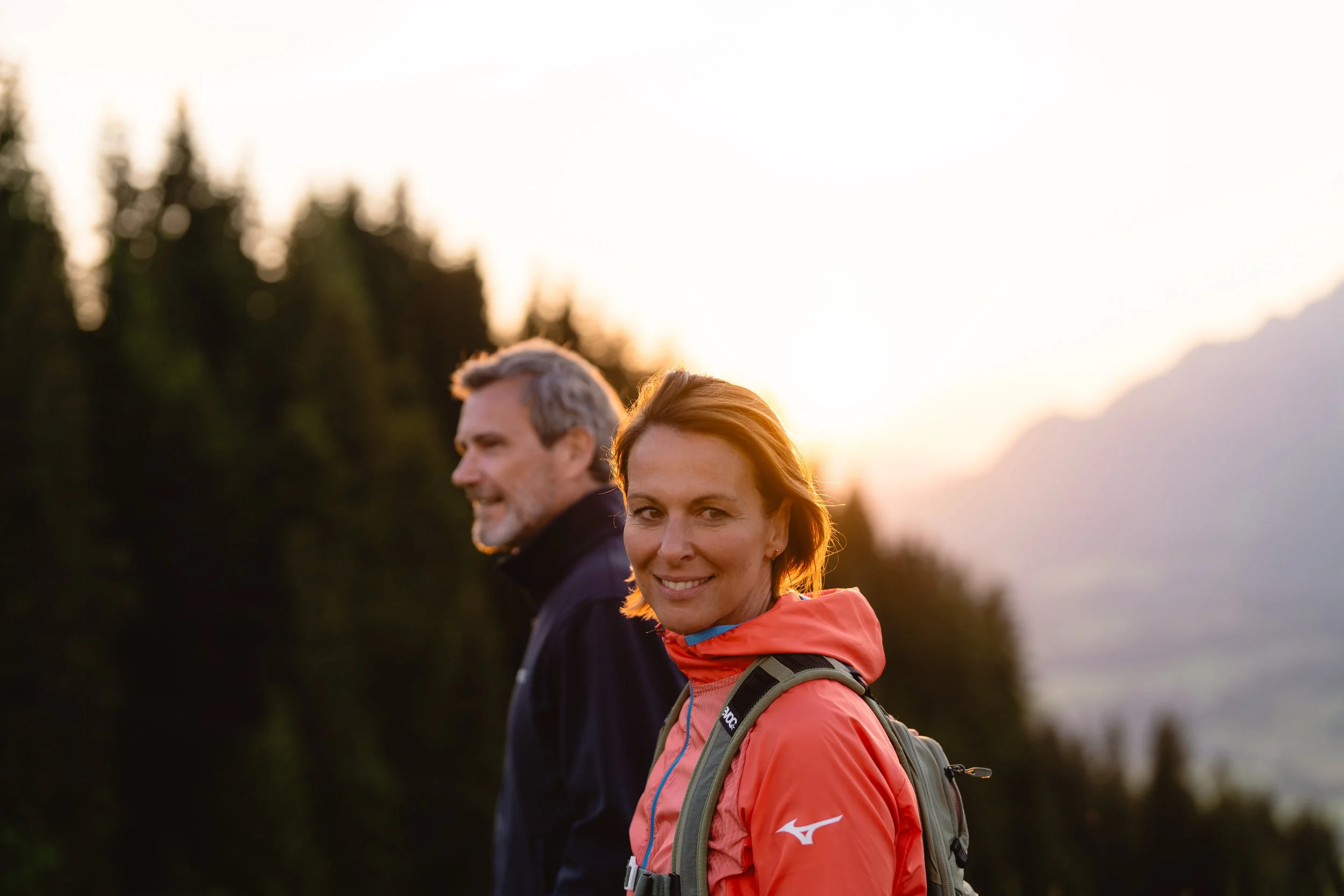 Ein Mann und eine Frau wandern in der Natur bei Sonnenuntergang, der Mann hat graue Haare und trägt eine dunkle Jacke, die Frau hat braune Haare, trägt eine rote Jacke und einen Rucksack.