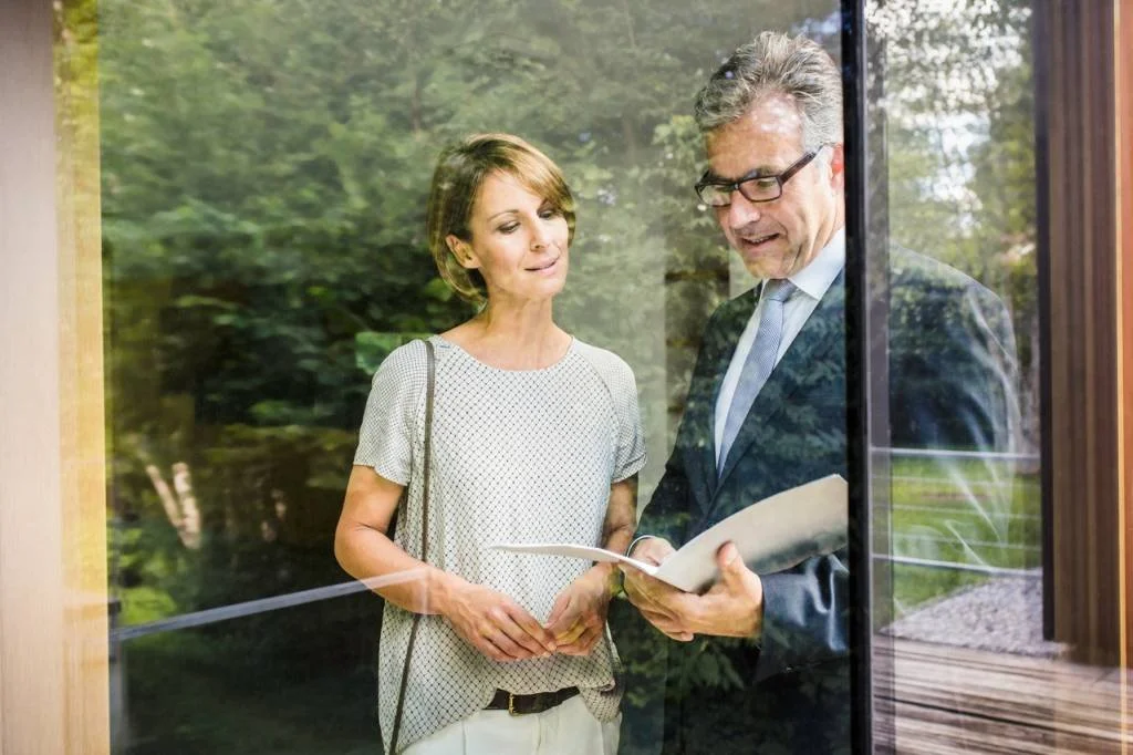 Ein Mann und eine Frau stehen bei einem Glaselement, der Mann liest aus einem Buch, die Frau schaut interessiert zu.