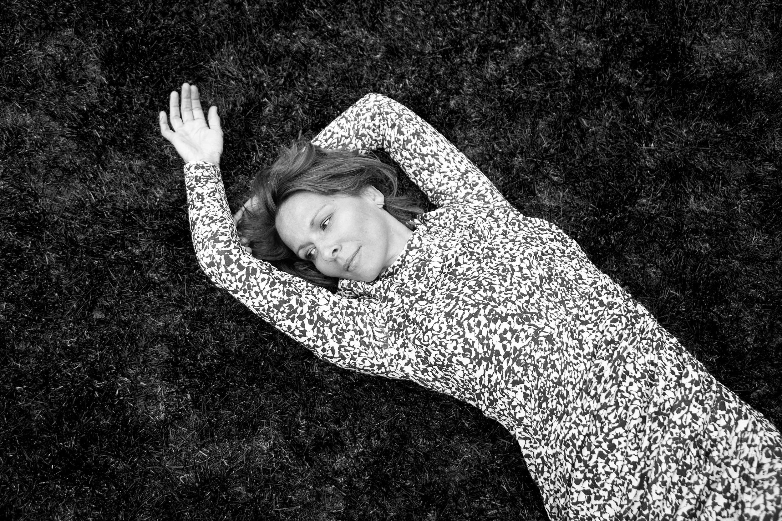 Eine Frau liegt entspannt auf dem Rücken auf einer Grasfläche, mit einem Muster-Kleid und den Armen über dem Kopf.