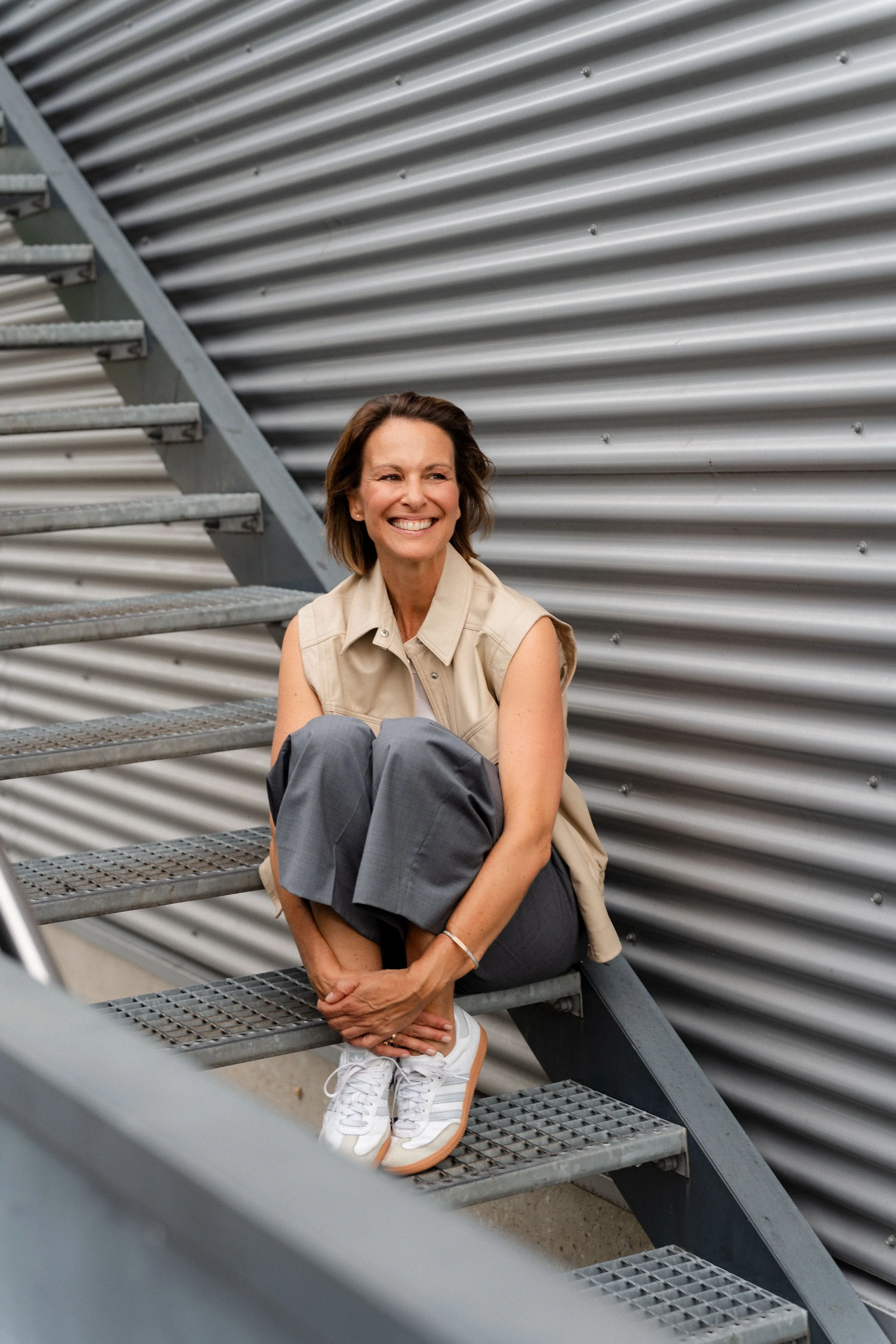 Junge Frau sitzt auf einer Metalltreppe vor einer silbernen, gewellten Wand, lächelt und trägt sportliche Kleidung.