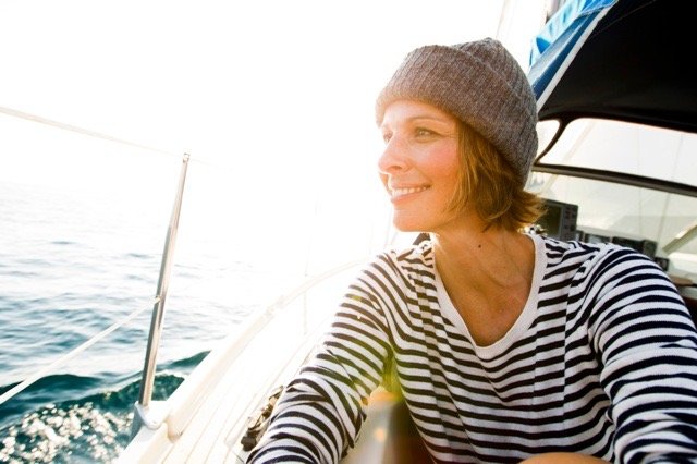 Frau mit gestreiftem Hemd und grauem Beanie auf einem Boot, blickt lachend in die Ferne, umgeben vom Meer und Sonne.