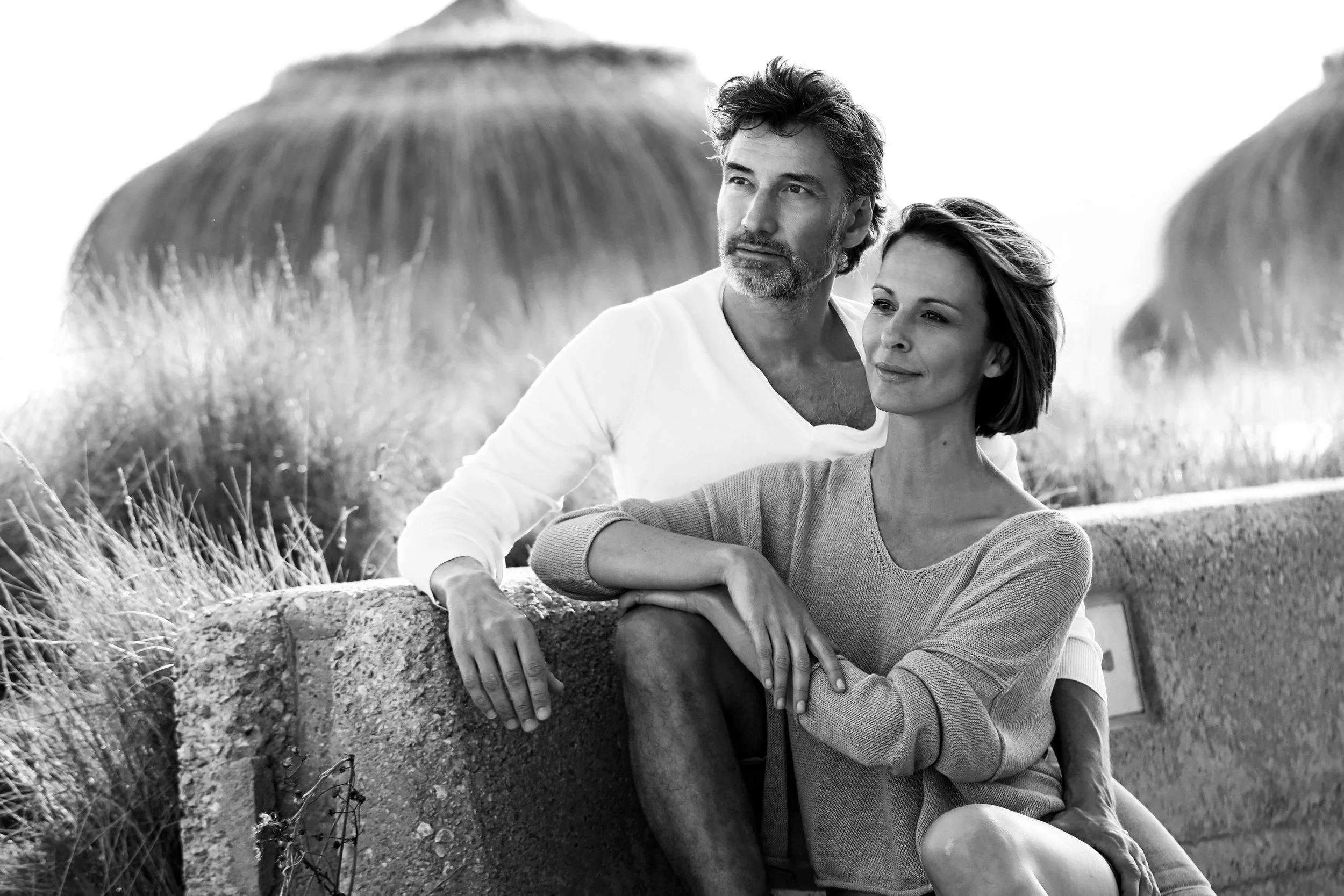 Ein Mann und eine Frau sitzen auf einer Mauer im Freien, im Hintergrund sind zwei große runde Hütten. Das Bild ist schwarz-weiß.