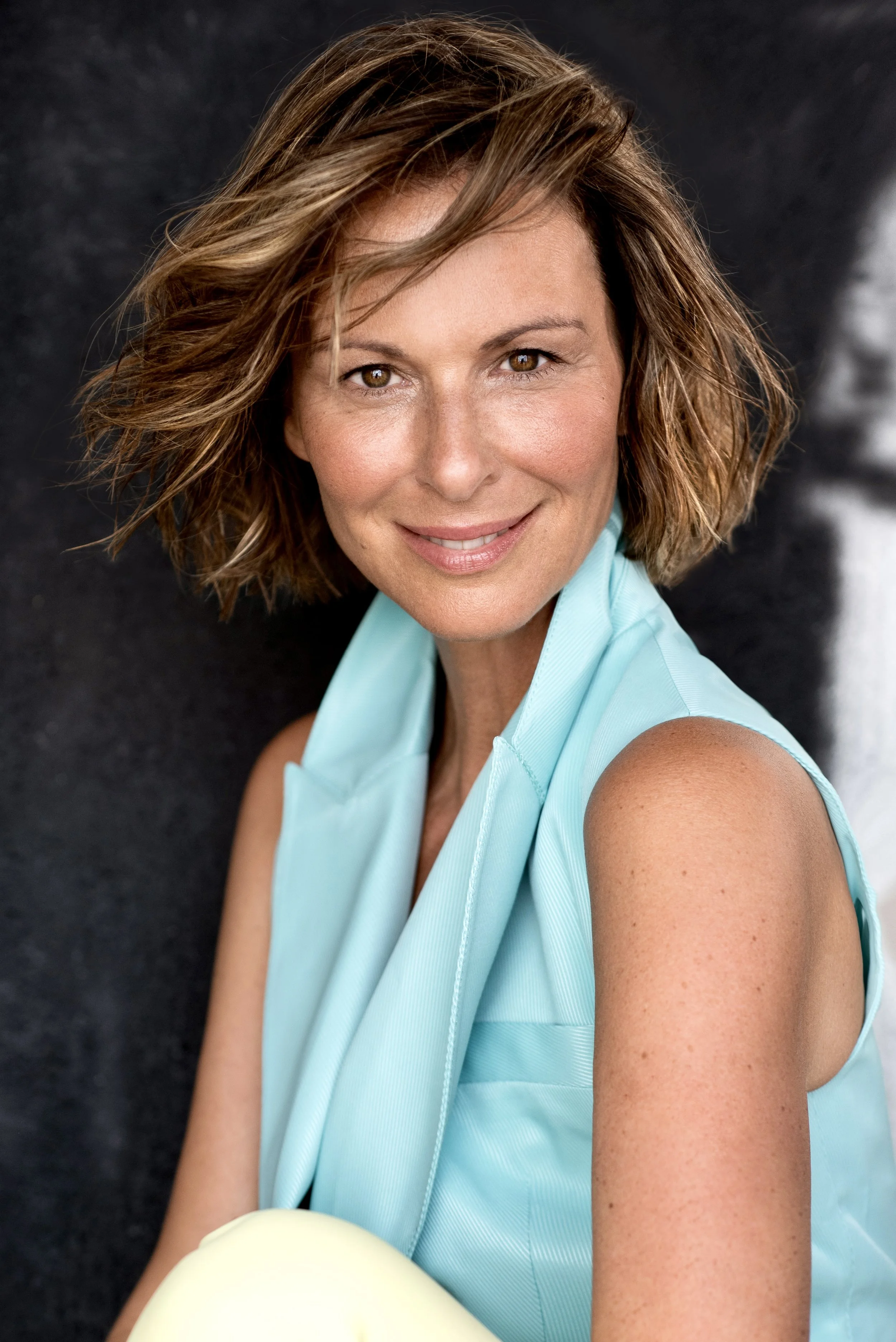 Eine Frau mit lockigem, schulterlangem braunem Haar trägt ein hellblaues Top und schaut lächelnd in die Kamera gegen einen dunklen Hintergrund.