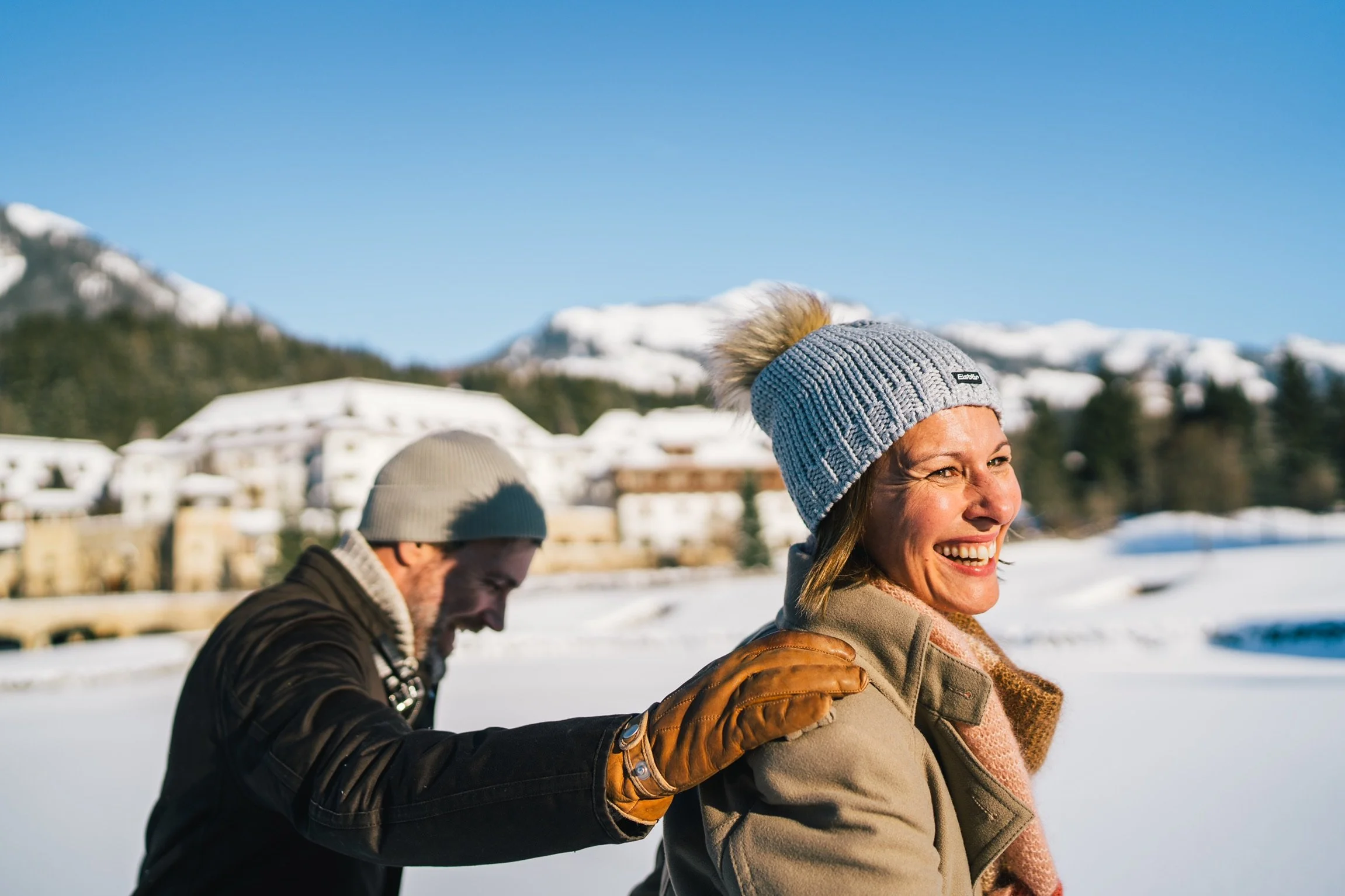 Ein lachendes Paar in Winterkleidung, mit Schnee bedeckte Berge im Hintergrund.
