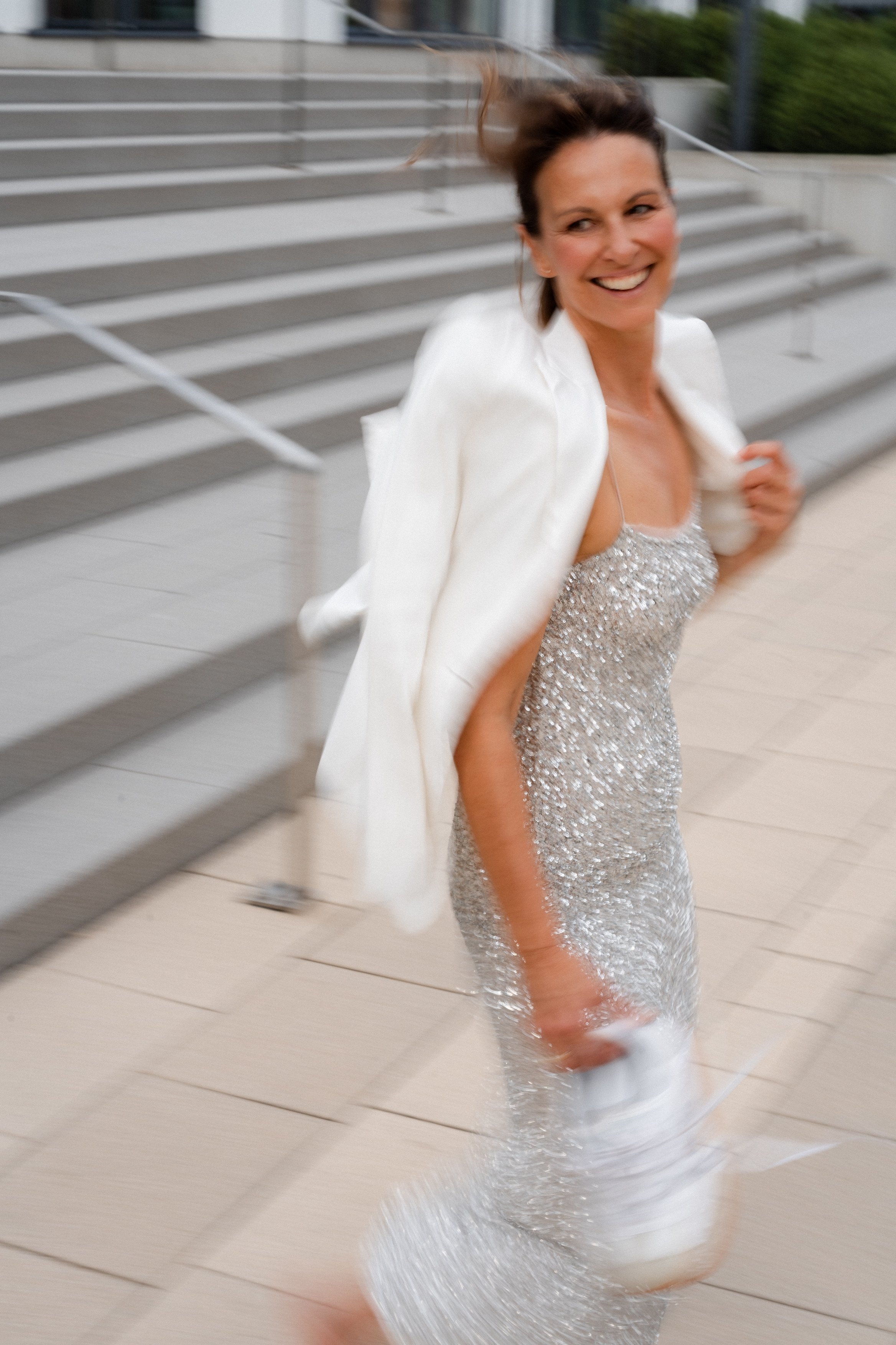 Eine lachende Frau in einem silbernen Pailletten-Kleid läuft auf einer Treppe. Sie trägt einen weißen Blazer über den Schultern.